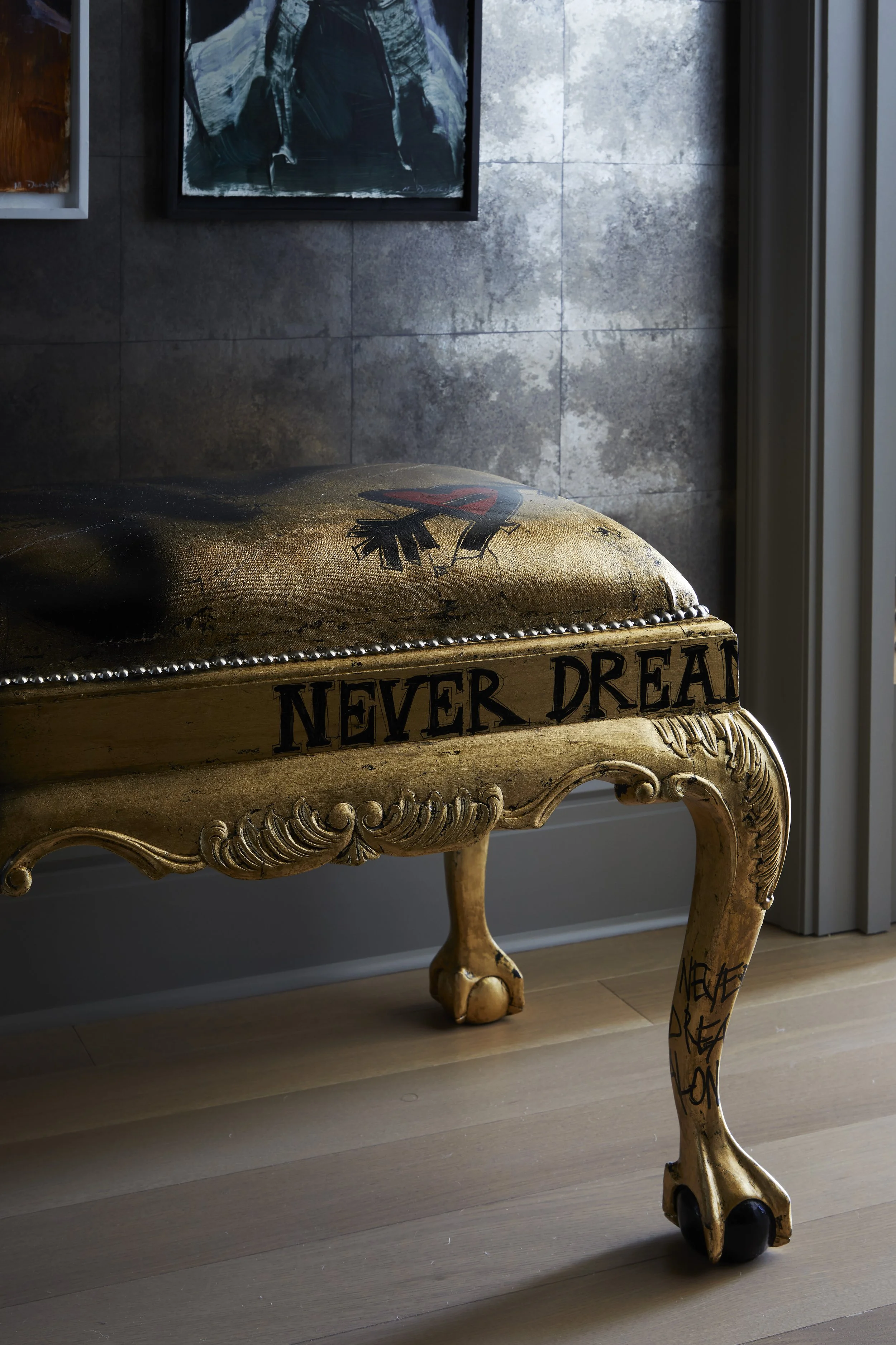 A vintage wooden bench with ornate carving and the words 'NEVER DREAM' painted on the front, situated near a wall with framed artwork, and a window or door with vertical blinds to the right.