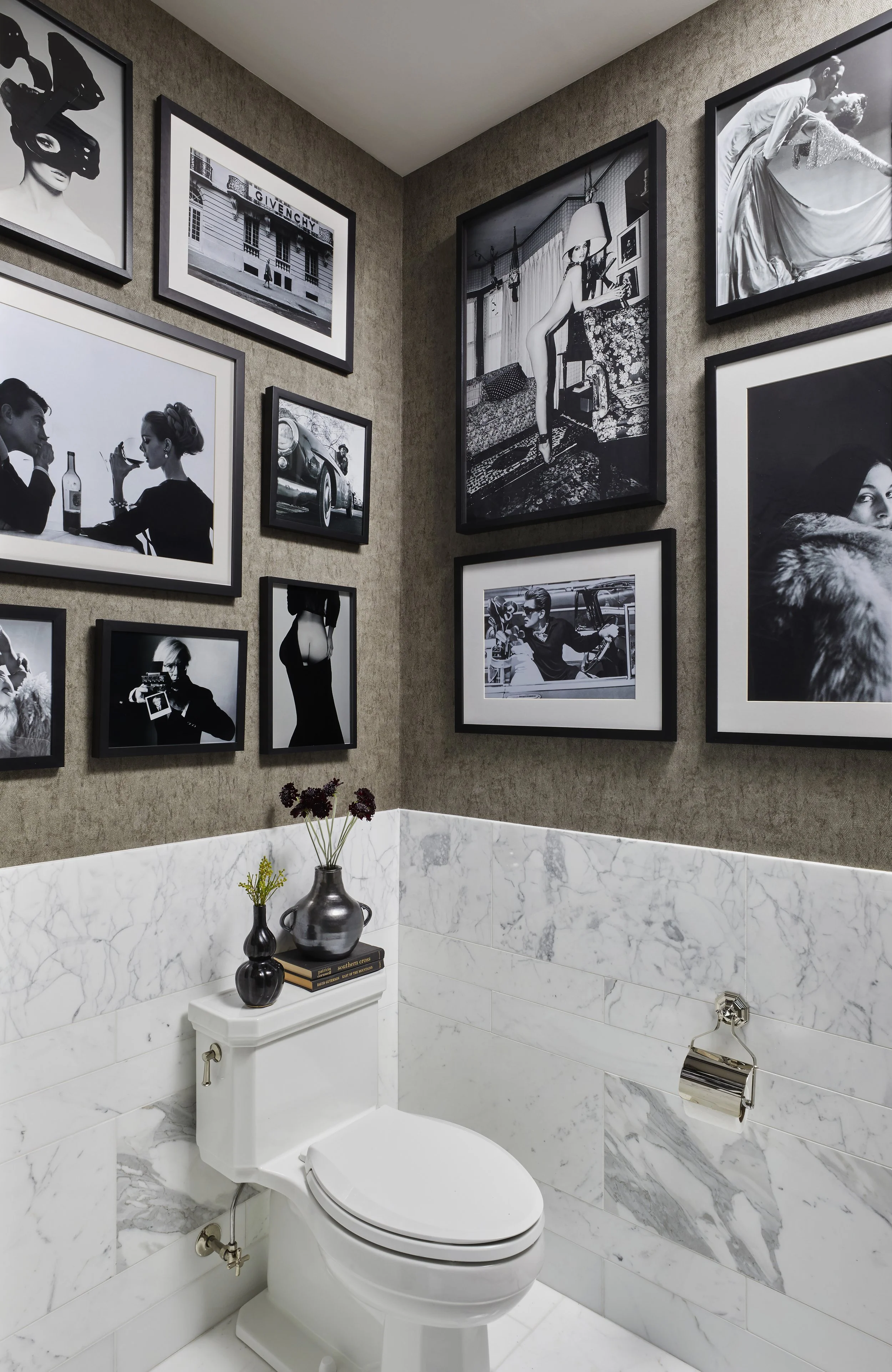 Bathroom with a modern white toilet, marble tile walls, and framed black and white photographs on the wall. A small white side table holds a metallic vase with purple flowers and a few books.