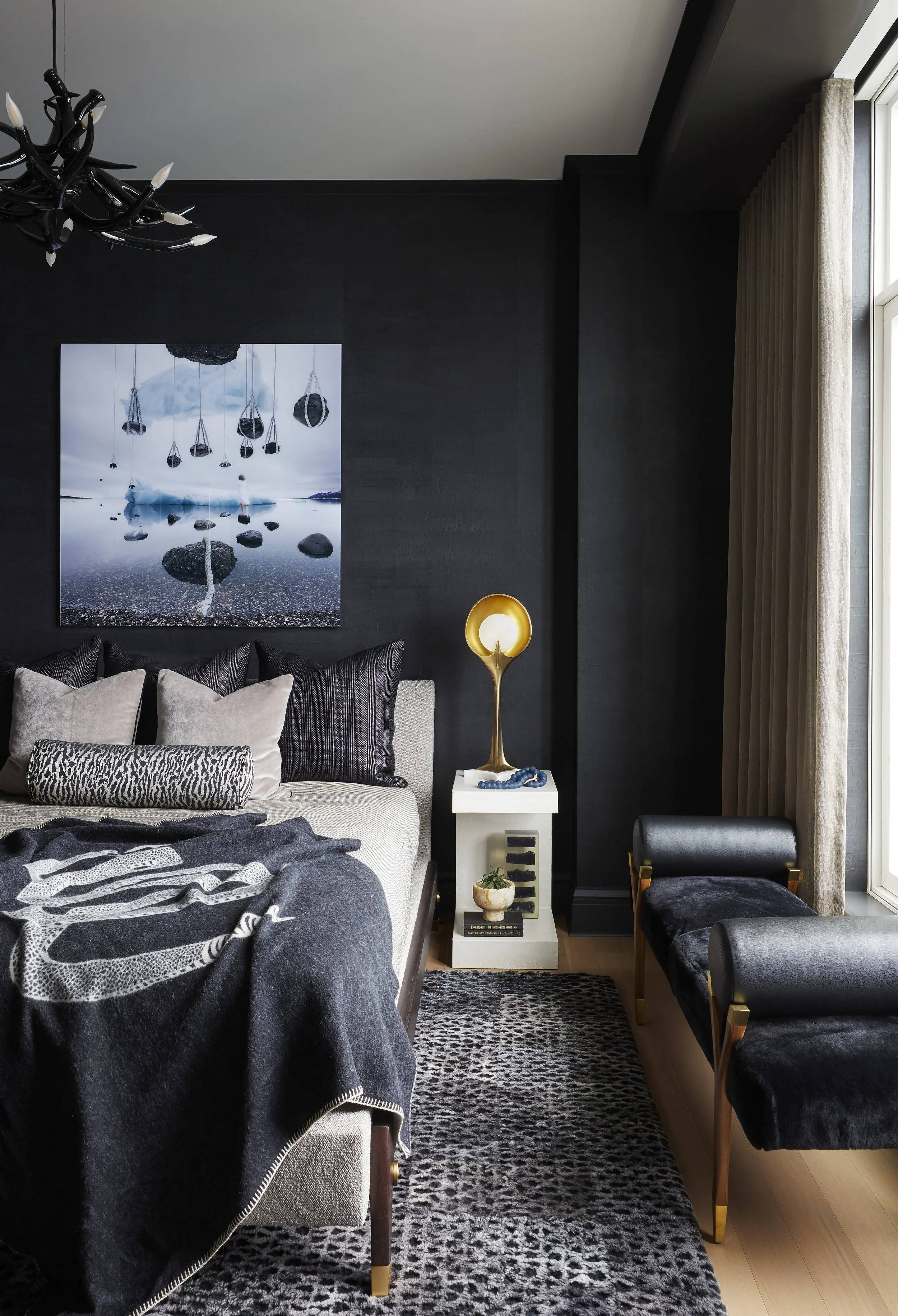 A modern living room with black walls, a white sofa with patterned and neutral pillows, a black and gold chair, and a patterned black and white rug. There's a white side table with a gold sculpture lamp, a small bowl, and books. A large abstract artw