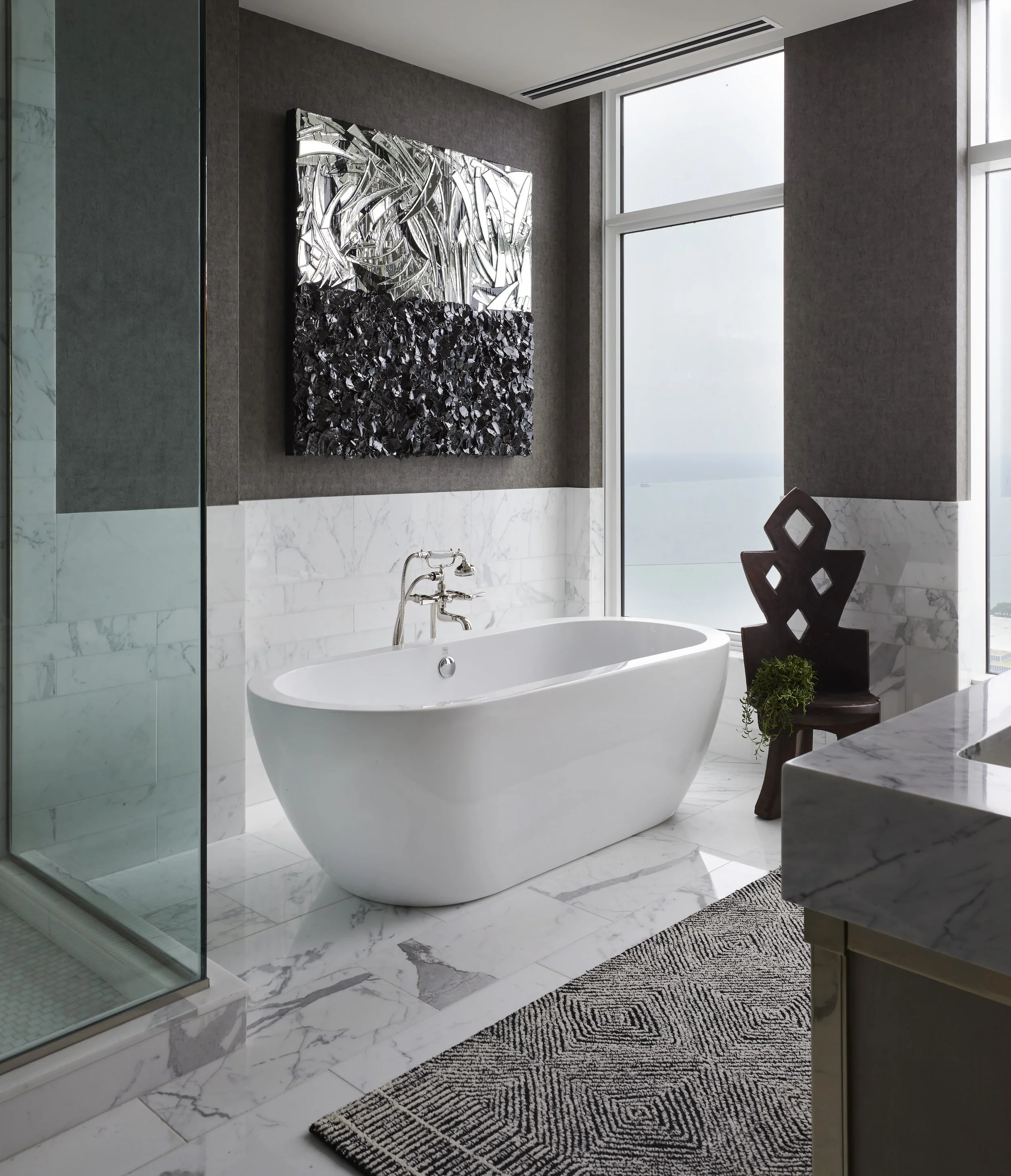 A modern bathroom with a freestanding white bathtub, a marble platform, and a large window. Decorative abstract metal wall art hangs above the tub. The bathroom has marble flooring, a glass shower enclosure, and a wooden chair with greenery, compleme