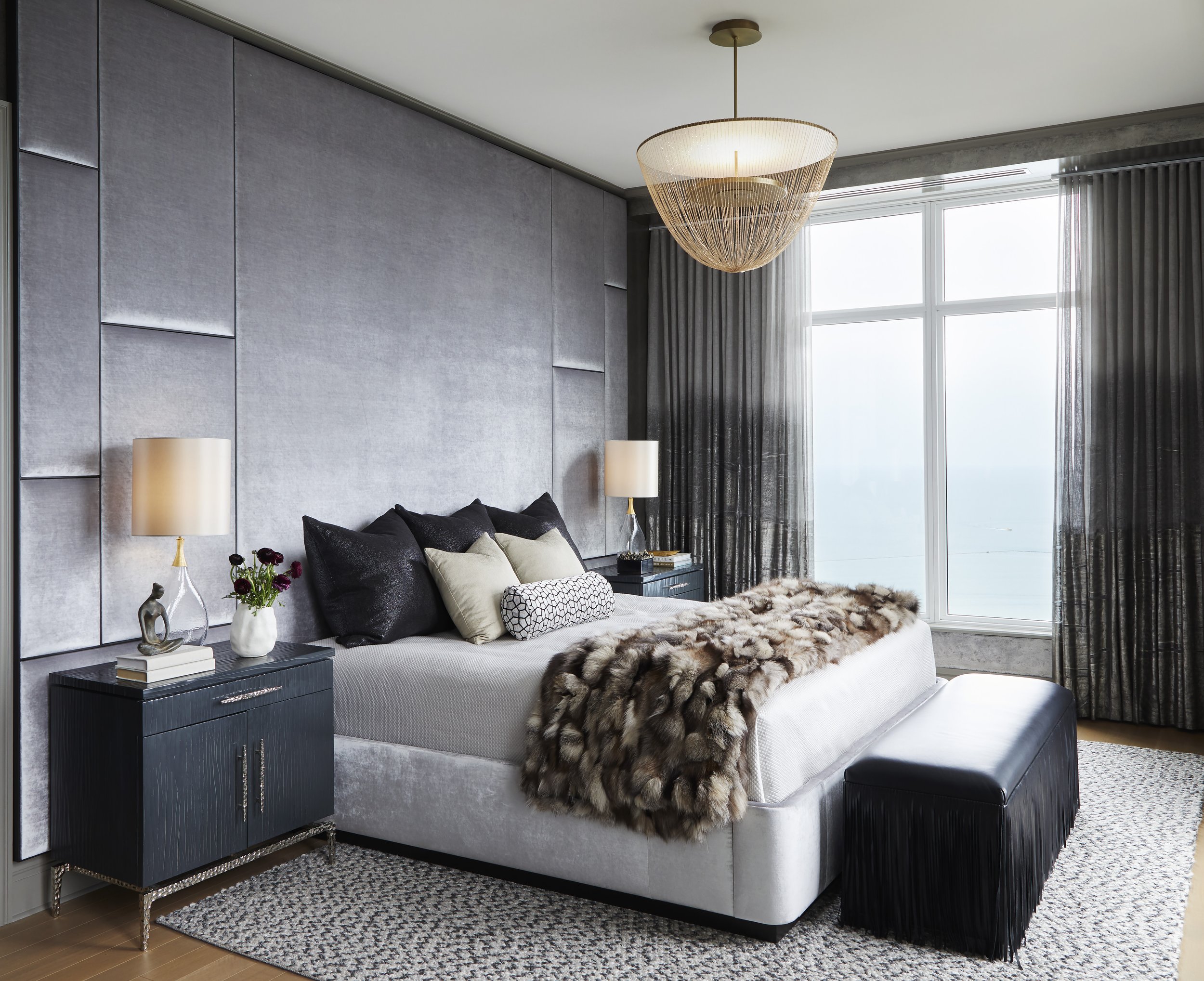 Living room with gray textured wall, large window with curtains, chandelier, black side table with books and sculptures, beige sofa with black, cream, and patterned pillows, faux fur throw, and black ottoman, over patterned rug on hardwood floor.
