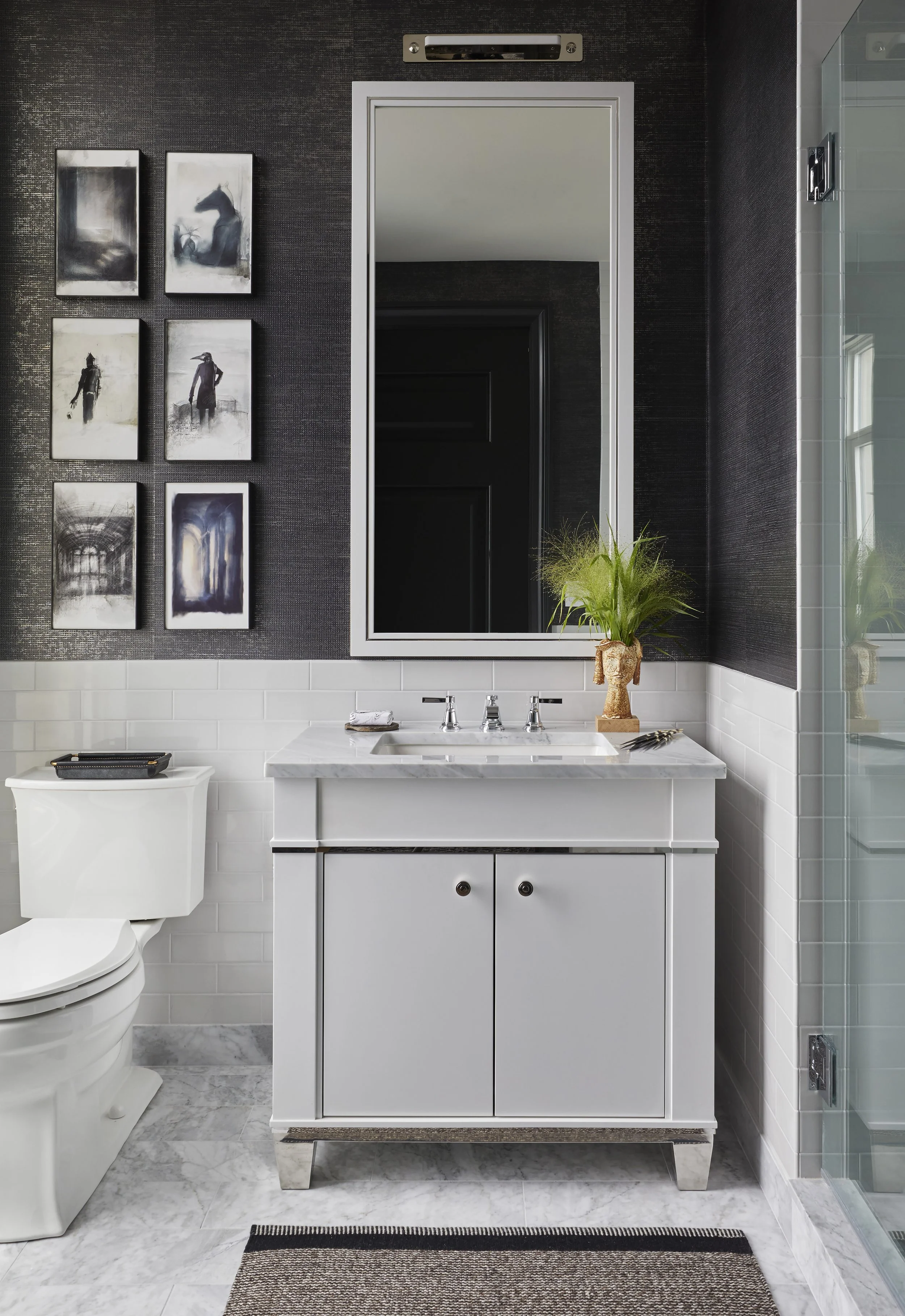 A modern bathroom with black textured wall paint, white tile below, a white vanity with a marble countertop, a white toilet, a large mirror, a small potted plant, and black-and-white artwork on the wall.