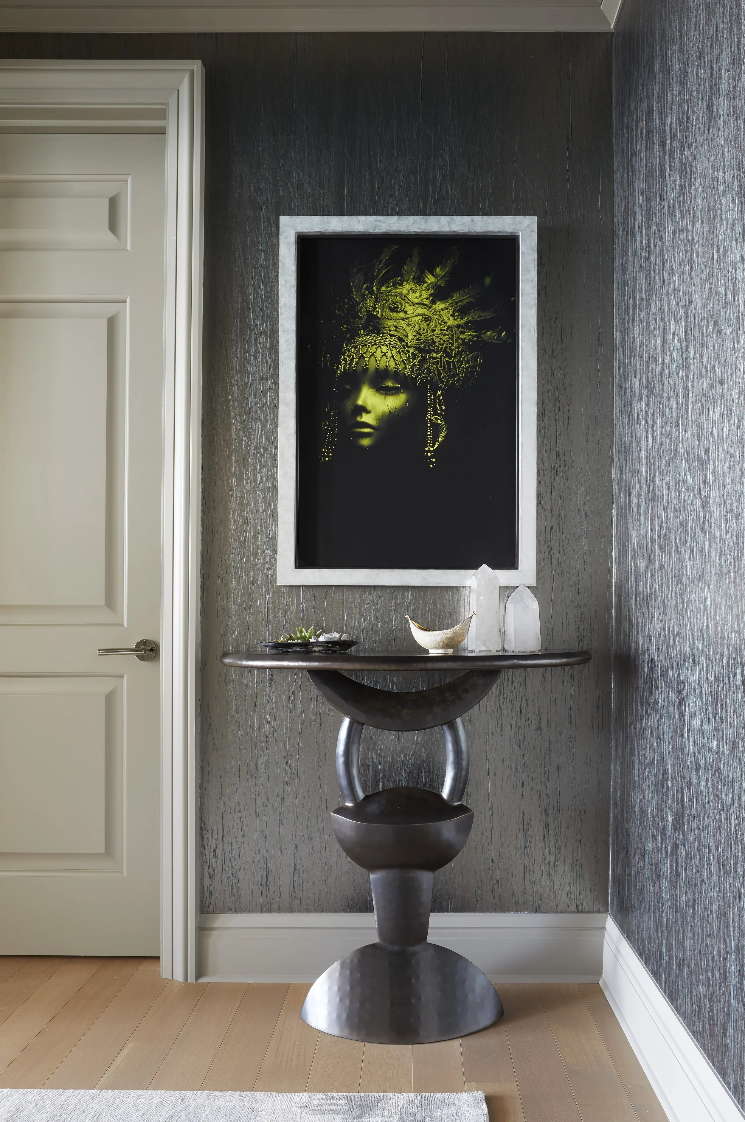 Interior view of a modern hallway showing a dark textured wall, a white door on the left, a framed artwork of a stylized face with an elaborate headdress, a small table with decor items, and a sculptural metal pedestal