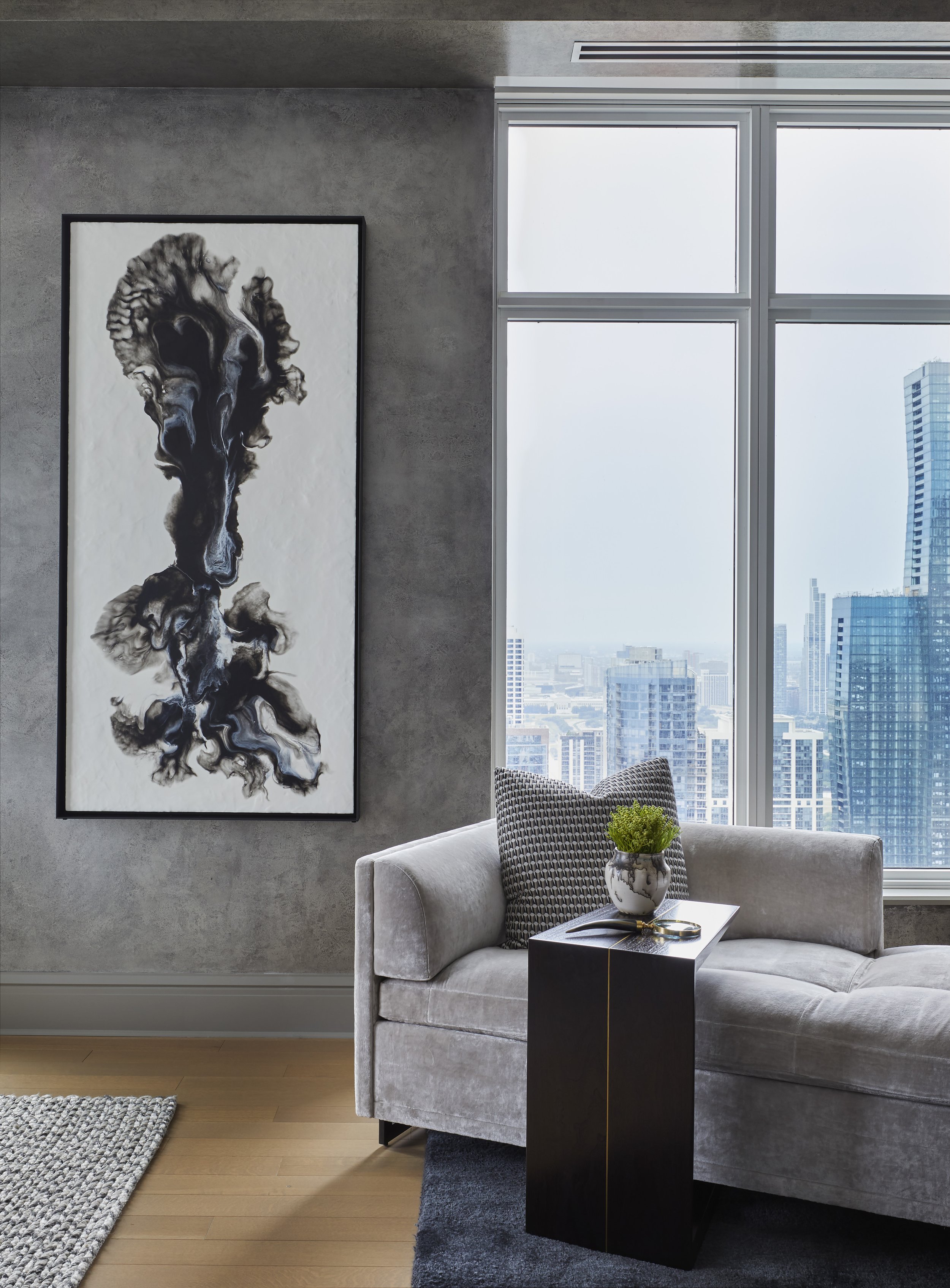 Modern living room with large window overlooking cityscape, gray sofa with black and white patterned pillows, black side table with plant in marble vase, abstract black and white art on gray wall, hardwood floor with textured rug.