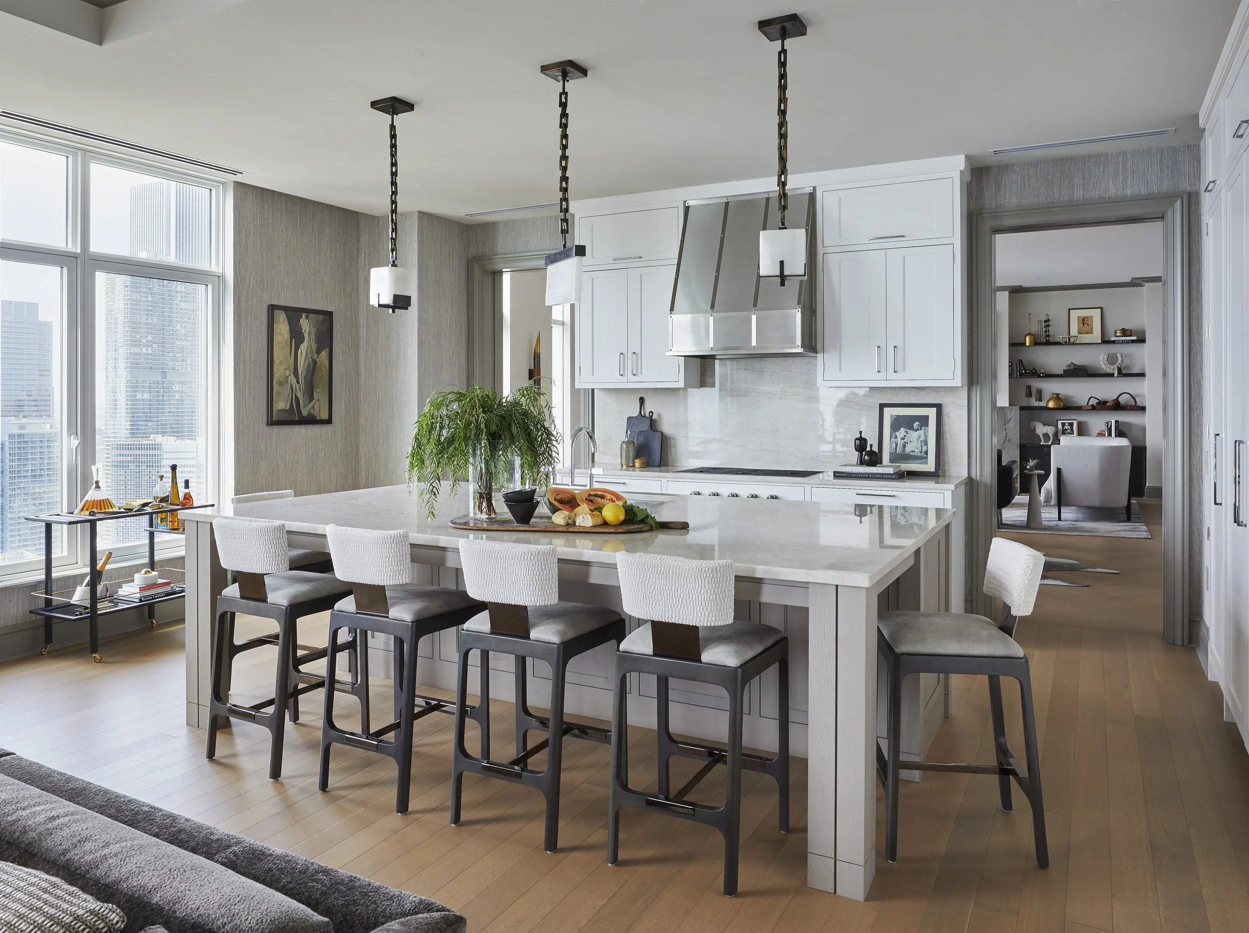 Modern kitchen with white cabinetry, a large white island with seating, pendant lighting, and large windows showing a city skyline.