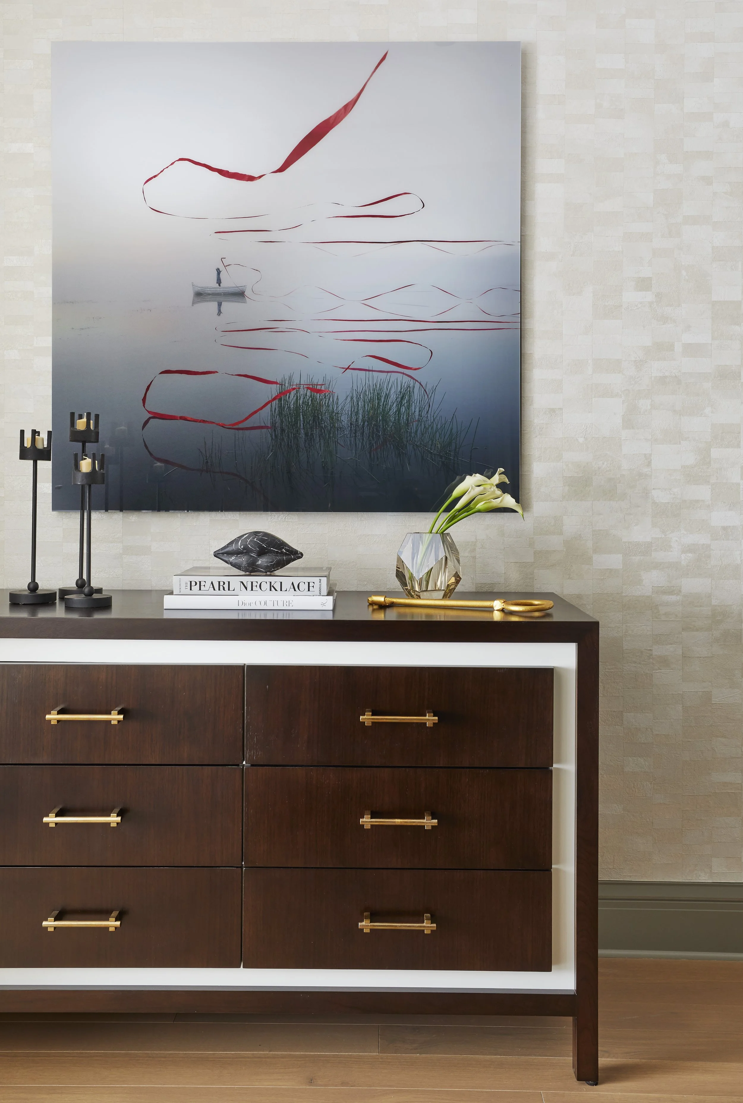 A wooden dresser with gold handles against a textured beige wall, topped with decorative items including black candle holders, a lapis lazuli stone, a white book titled 'PEARL NECKLACE', a vase with white calla lilies, and a pair of gold scissors. Ha