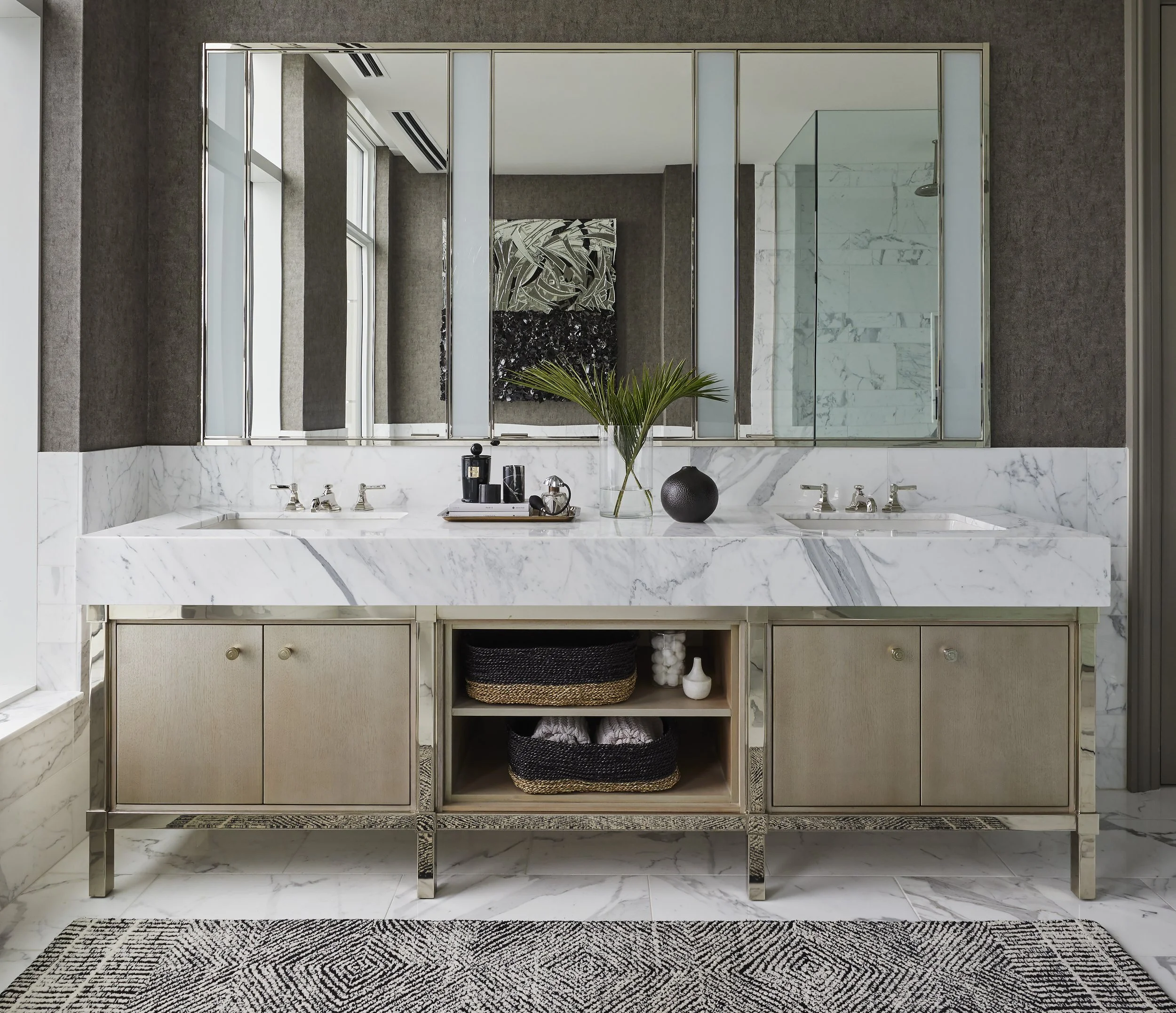 Modern bathroom vanity with a marble countertop, a large mirror, and storage shelves underneath, decorated with a black vase, a tray with toiletries, and a black diffuser, with a striped rug on the marble floor.