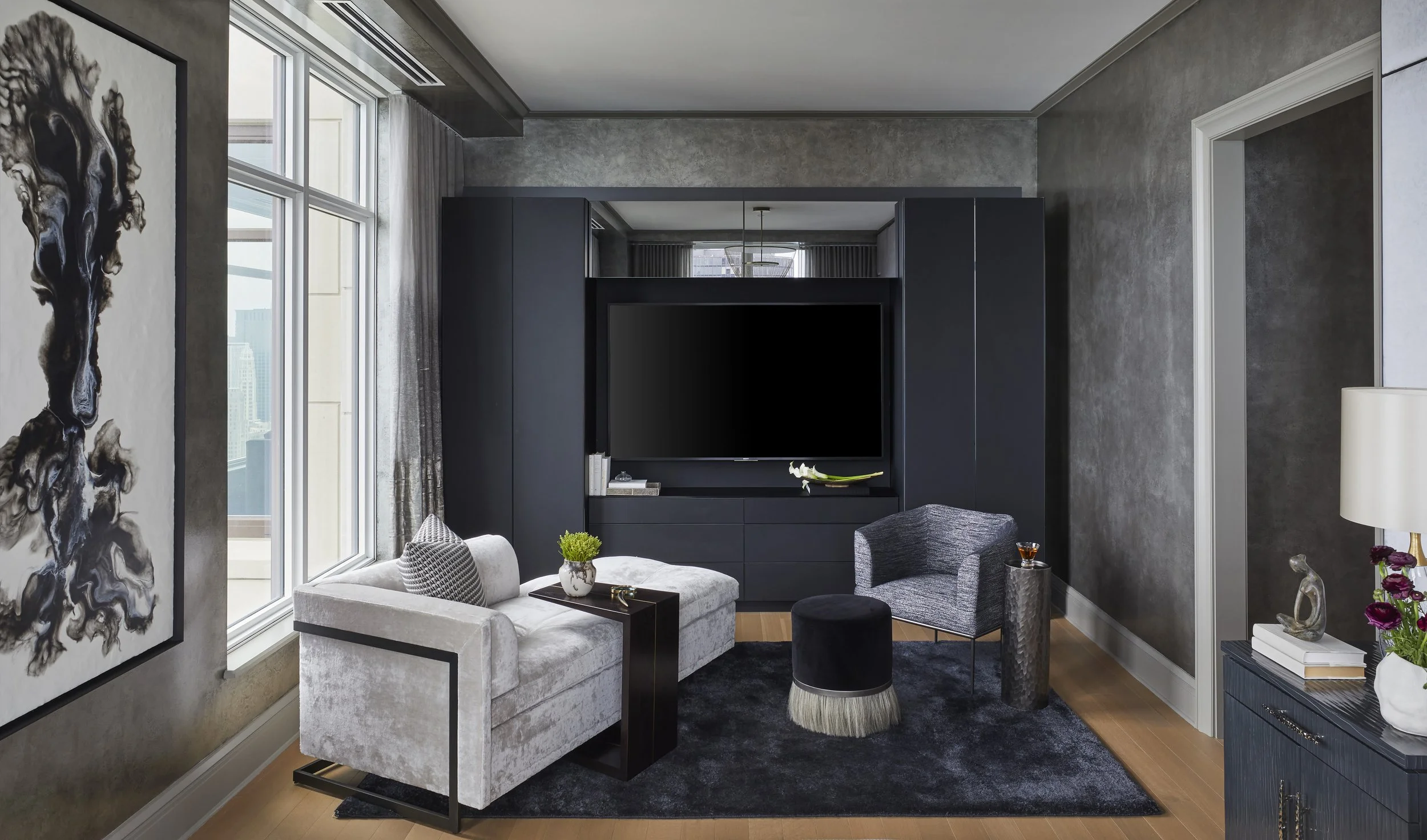 A modern living room with a large window, gray walls, a black entertainment center with a flat-screen TV, and various furniture including a white couch, gray chairs, and decorative plants and artwork.