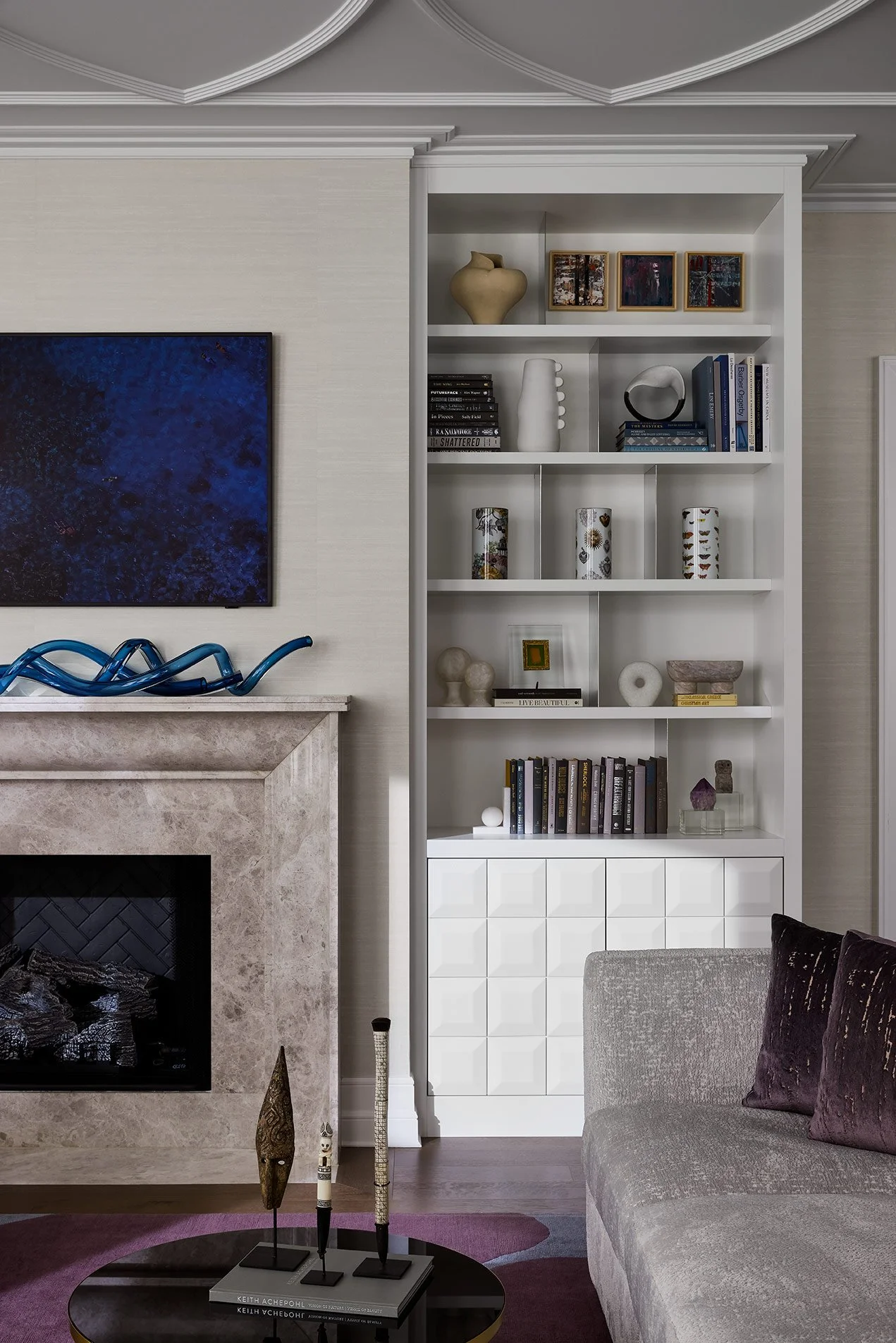 A living room corner with a marble fireplace, a gray sofa with a dark purple pillow, a black coffee table with decorative objects, a white bookshelf filled with books and decorative items, and a wall-mounted TV. The ceiling has decorative molding.
