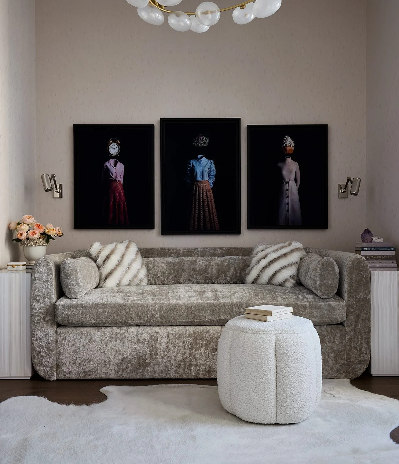 Living room with a beige velvet sofa, fur pillows, a white ottoman with books, a white furry rug, wall art featuring vintage women, a flower vase with roses, and modern wall lamps.