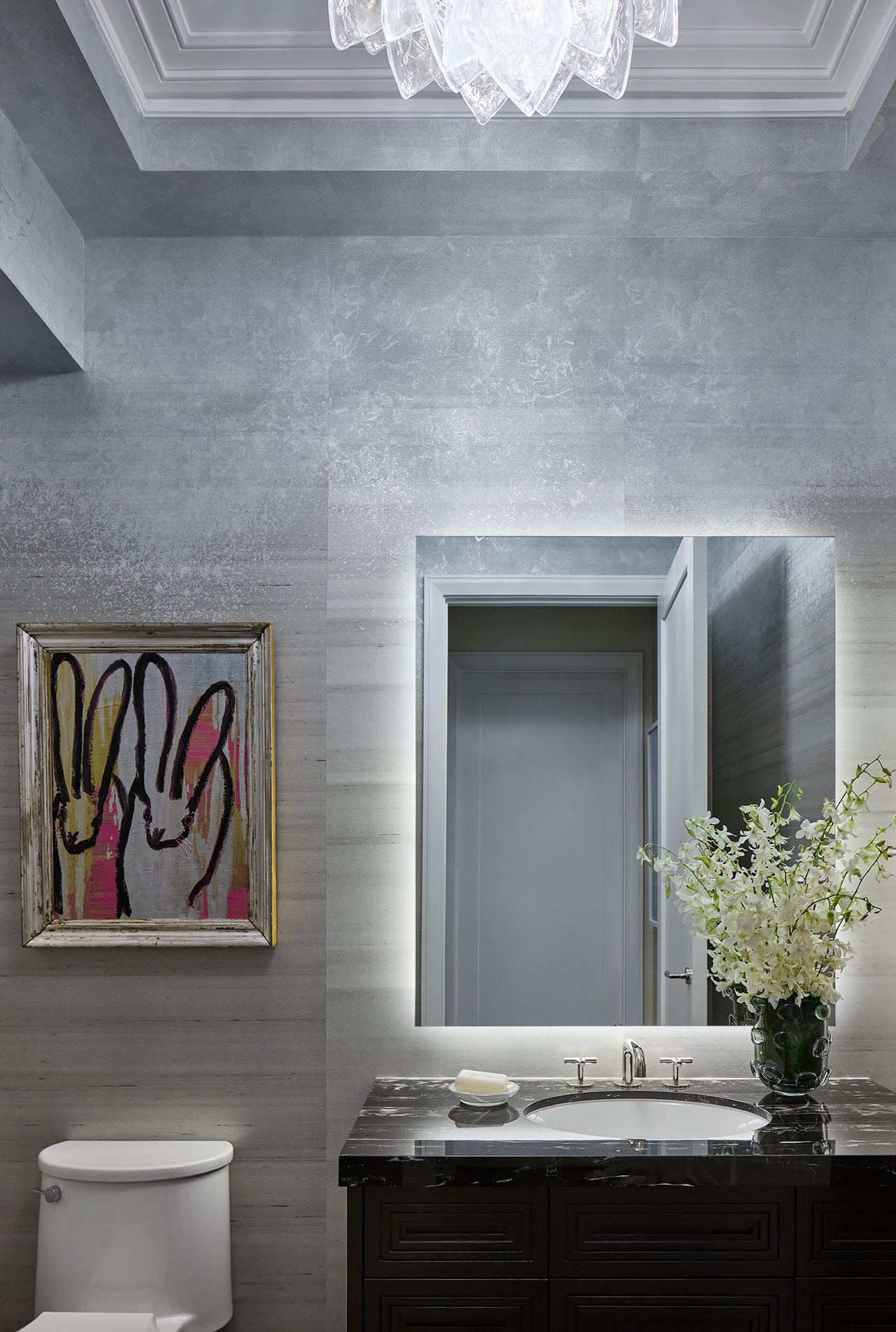 Modern bathroom with a large rectangular mirror, black marble sink countertop, white vase with white flowers, artwork on the wall, and a white toilet.