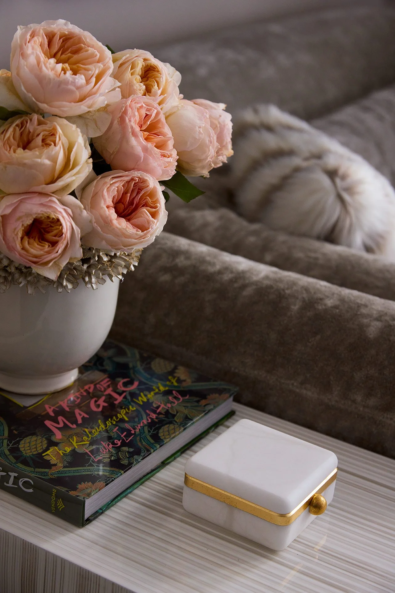 A white vase filled with pink and cream-colored roses is placed on a white surface. Nearby, there is a colorful book titled 'The Kaleidoscope of Woman'. Also on the surface, a white rectangular box with a gold band and a small gold sphere is visible.