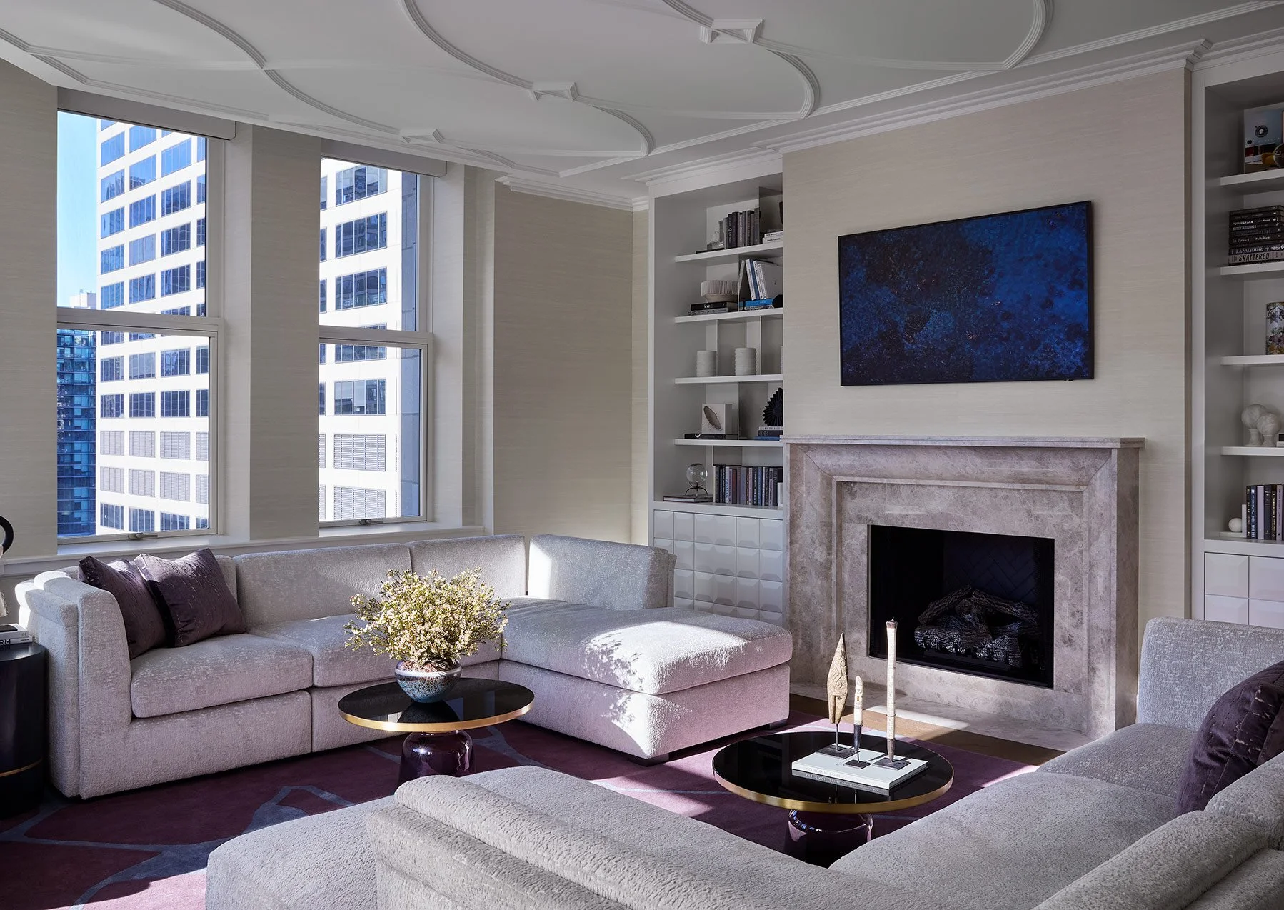 Living room with large windows showing tall modern buildings outside, cream-colored sofas, a pink and purple area rug, black coffee tables with decorative items, a fireplace with a pink marble mantel, a wall-mounted TV, built-in shelves with books an