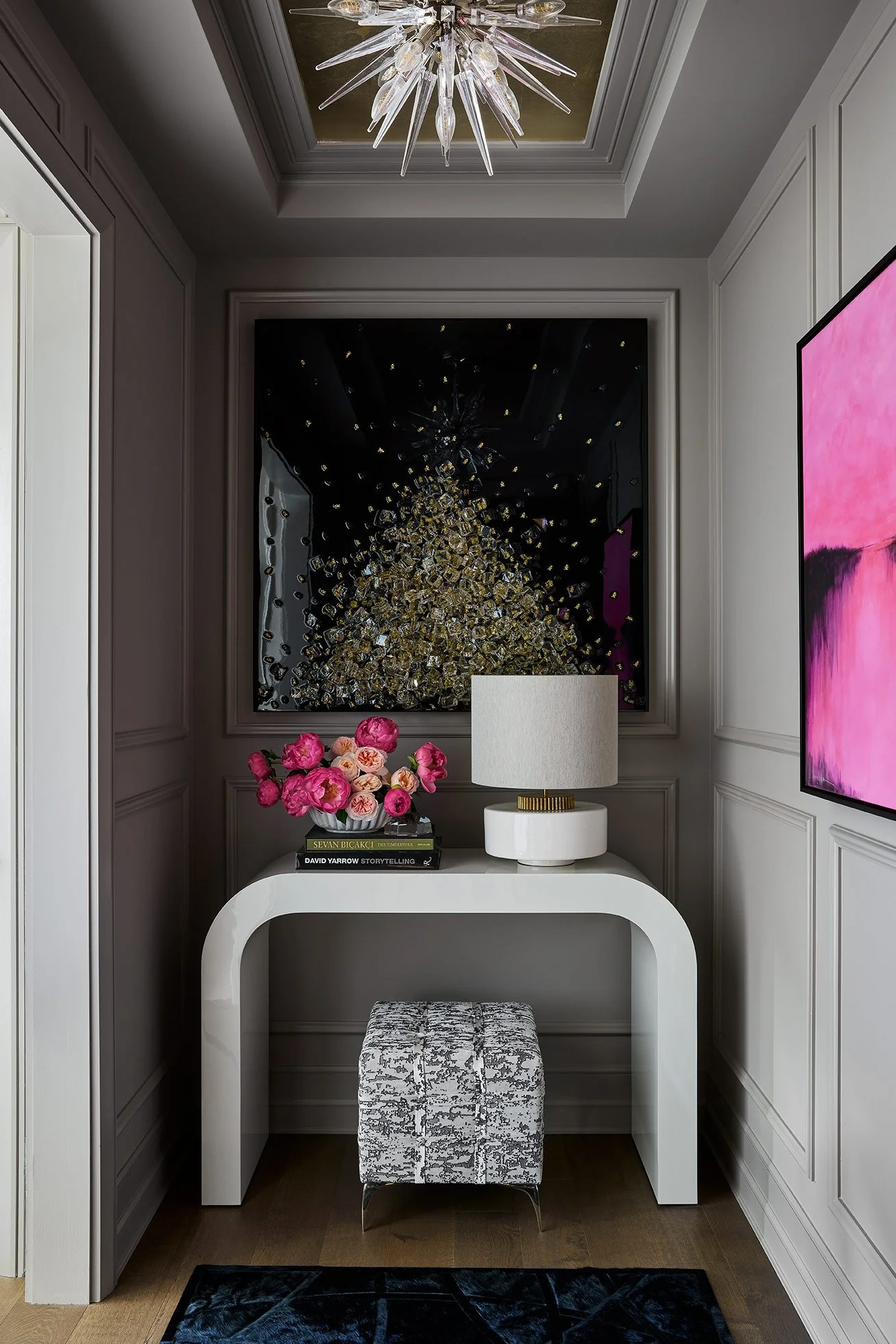 A stylish hallway corner with a white console table, a marble-patterned stool, a bouquet of pink flowers, a white table lamp, a large black artwork with gold details, a modern chandelier on the ceiling, and a pink abstract painting on the wall.
