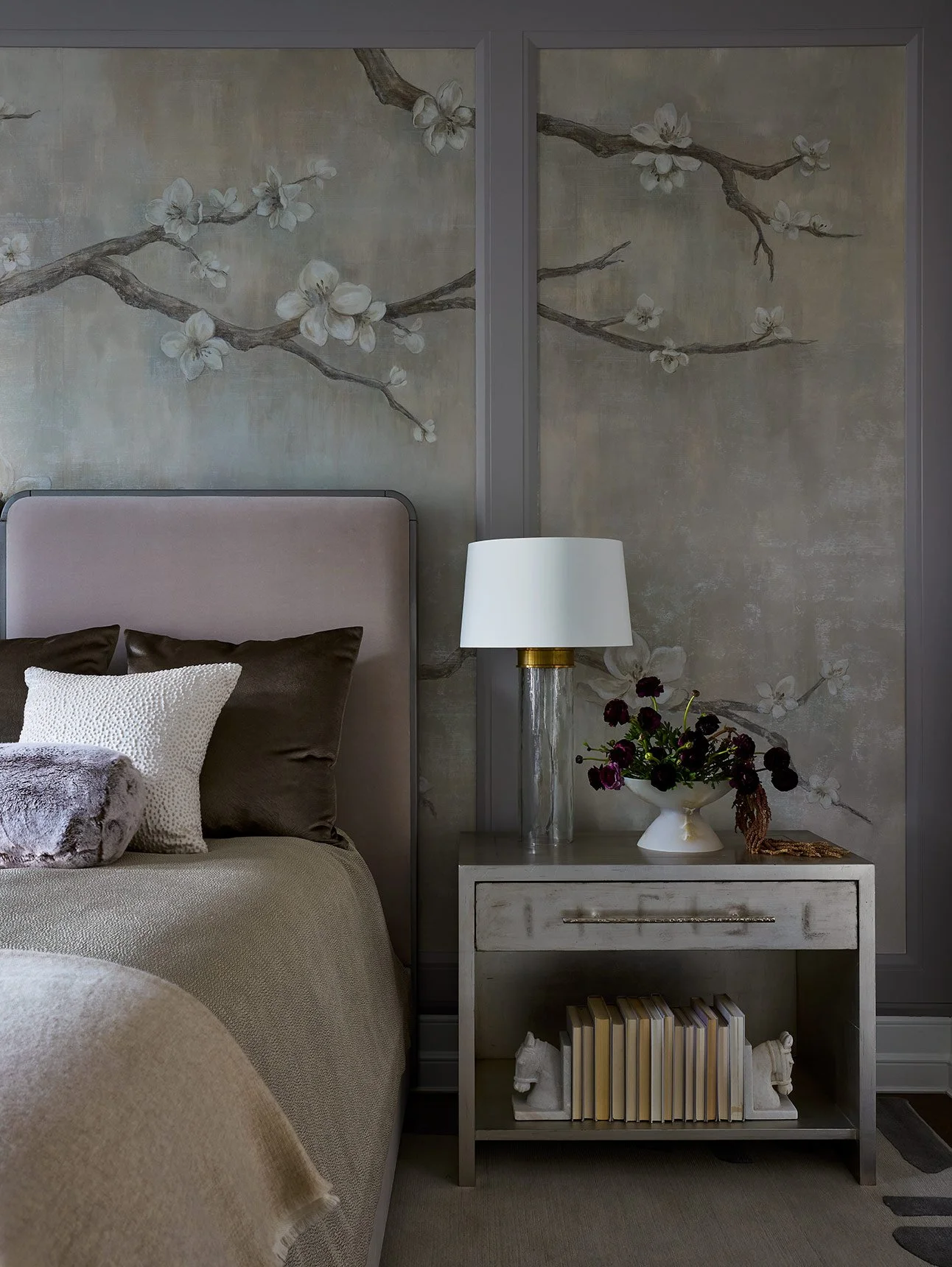 A bedroom with a beige bed, dark brown pillows, and a white pillow. A nightstand with a white lamp and a vase of dark purple flowers sit next to the bed, with framed floral artwork on the wall behind.