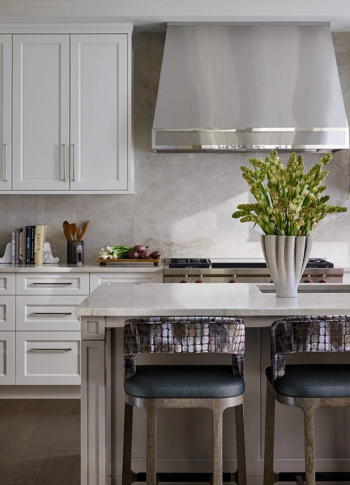 A kitchen with white cabinets, a marble countertop, a large vase with white flowers, a stove, and a variety of cooking utensils and vegetables on the counter.