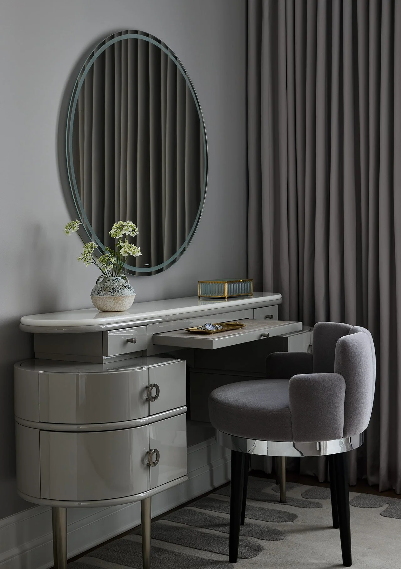 Elegant vanity table with a large oval mirror, decorated with a vase of white flowers, and a gray upholstered chair, set against gray curtains and a striped rug.