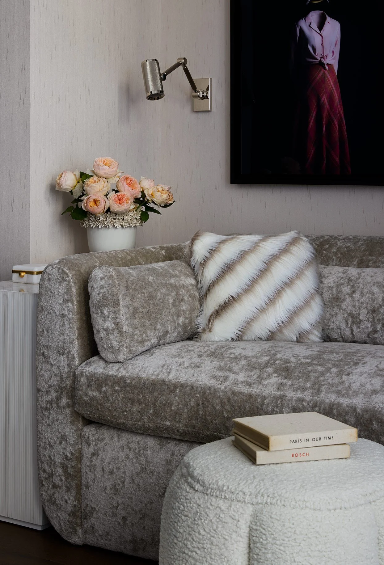 A plush velvet sofa with a decorative striped faux fur pillow, a small white round ottoman with books on it, a bouquet of pink peonies in a white vase, a wall-mounted adjustable reading light, a framed photograph of a woman in a tied blouse and plaid