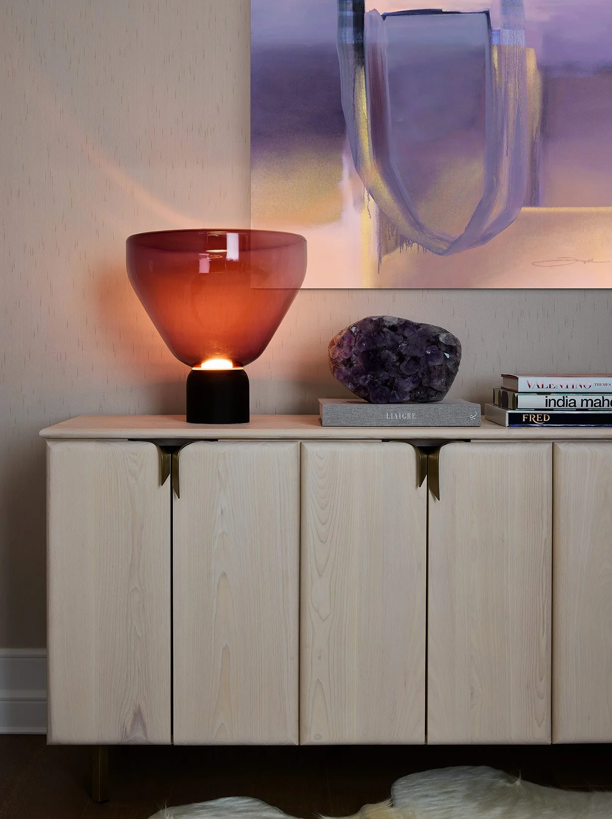A wooden sideboard with a red glass lamp, a purple gemstone sculpture on a gray book, and a stack of books on top. Abstract artwork hanging on the wall above.