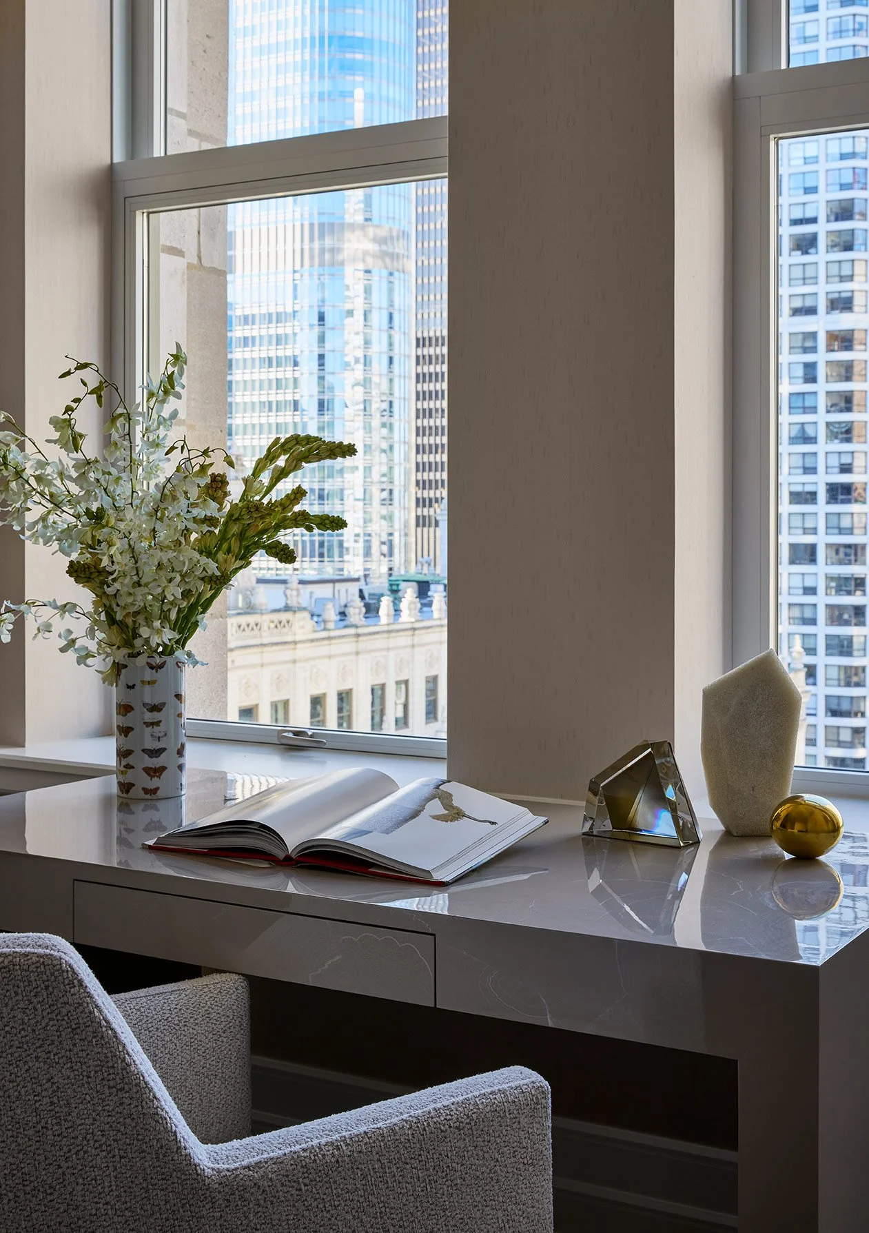 A desk with a large open book, a vase of white flowers, a decorative sculpture, and a small golden object in front of big windows showing tall modern buildings outside.