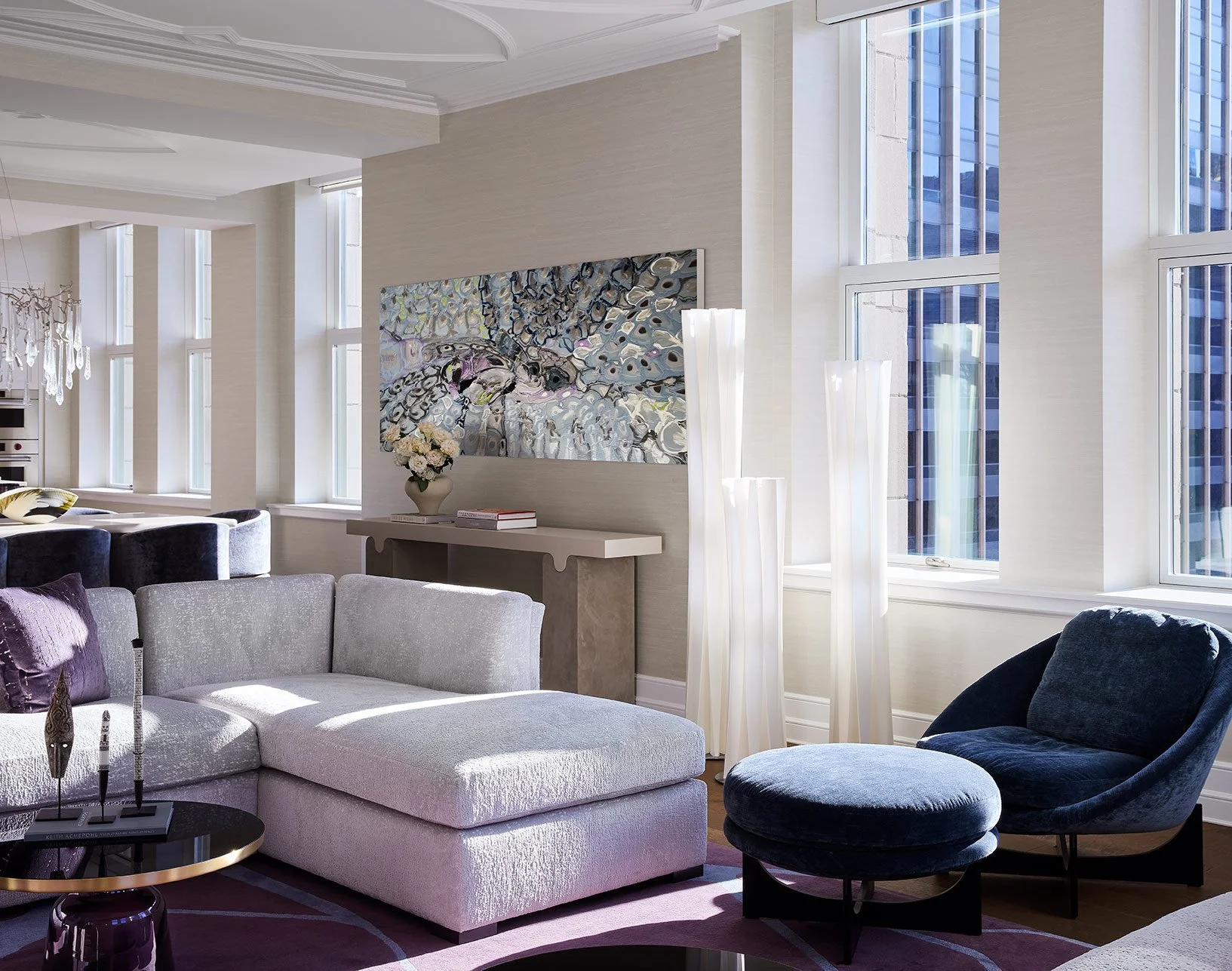 Bright living room with large windows, white walls, modern artwork, and contemporary furniture including a sectional sofa, lamp, and decorative accents.