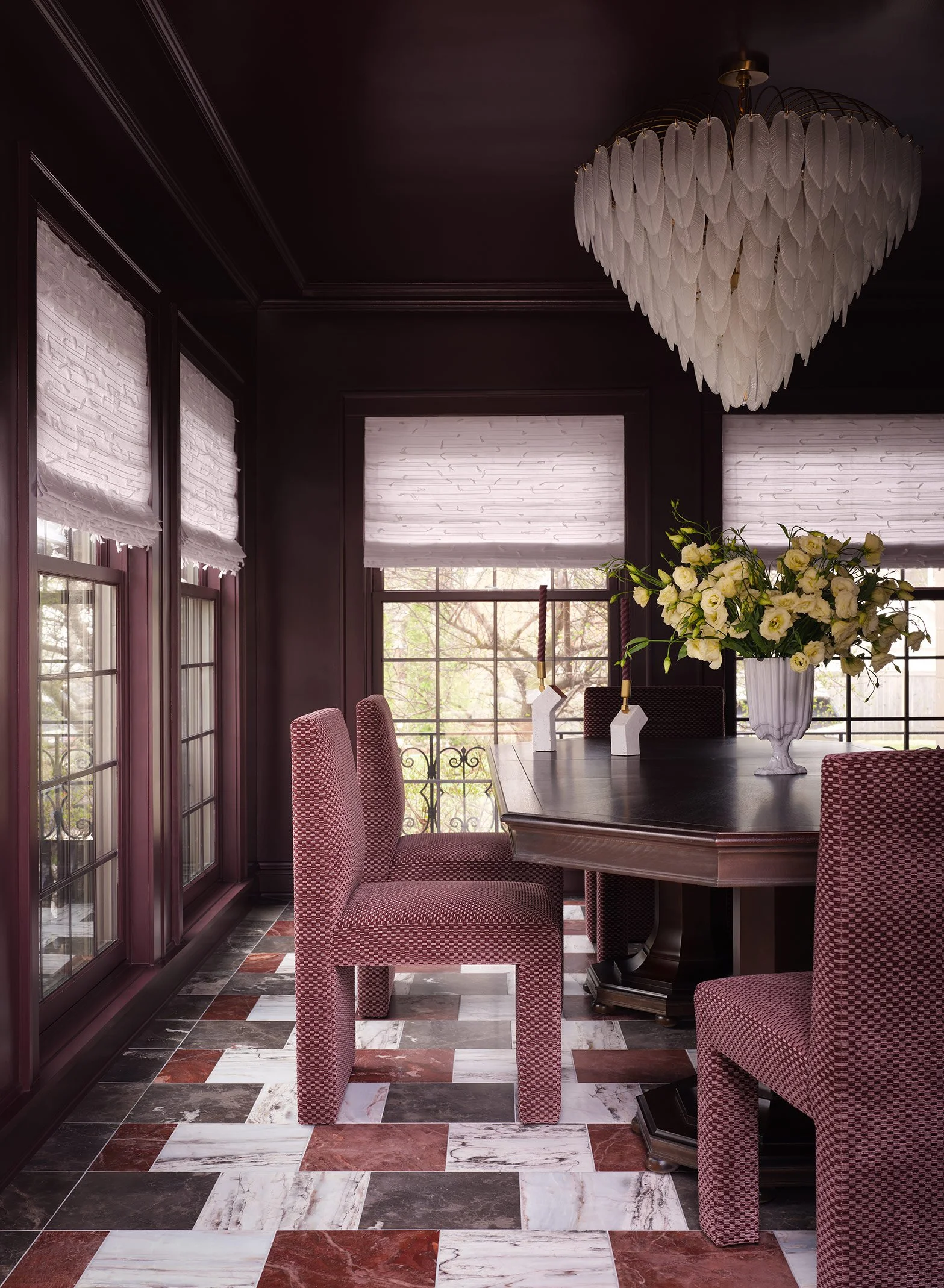 Dining room with a dark ceiling, white window shades, a large chandelier made of white feathers in the shape of a heart, a dark dining table with a floral centerpiece, and pink upholstered chairs with a diamond pattern.