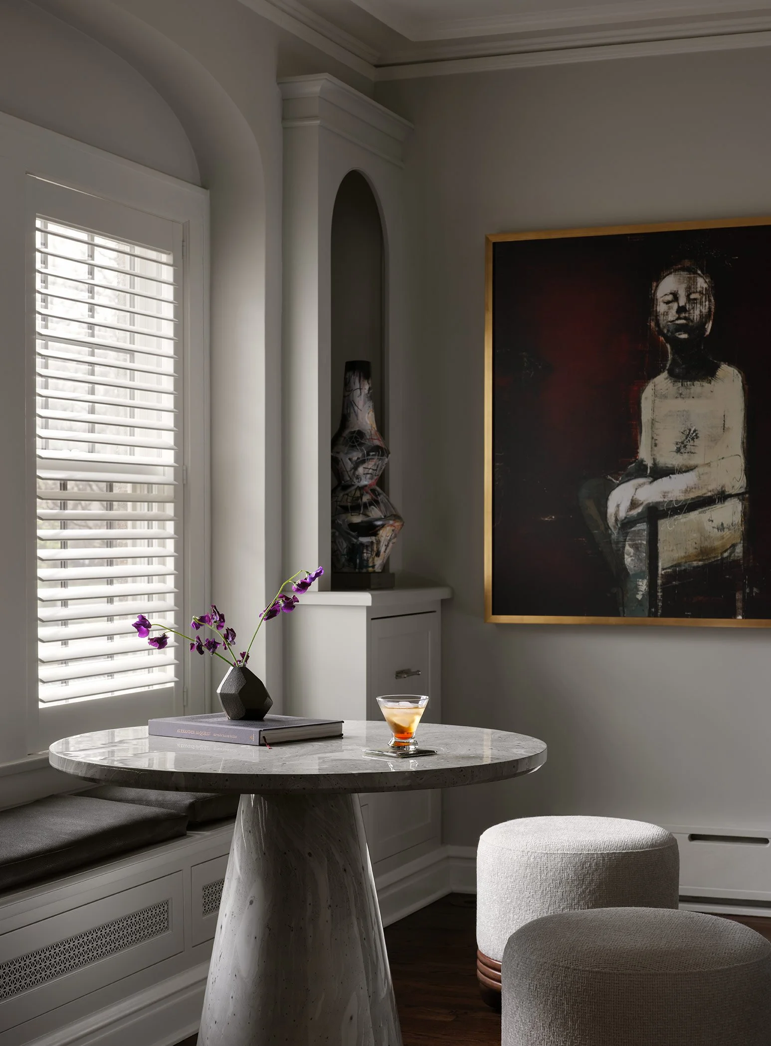 A cozy interior scene featuring a marble dining table with a dark geometric vase holding purple flowers, a glass of whiskey, a large framed painting of a seated woman, window with white plantation shutters, and two round upholstered stools in a livin