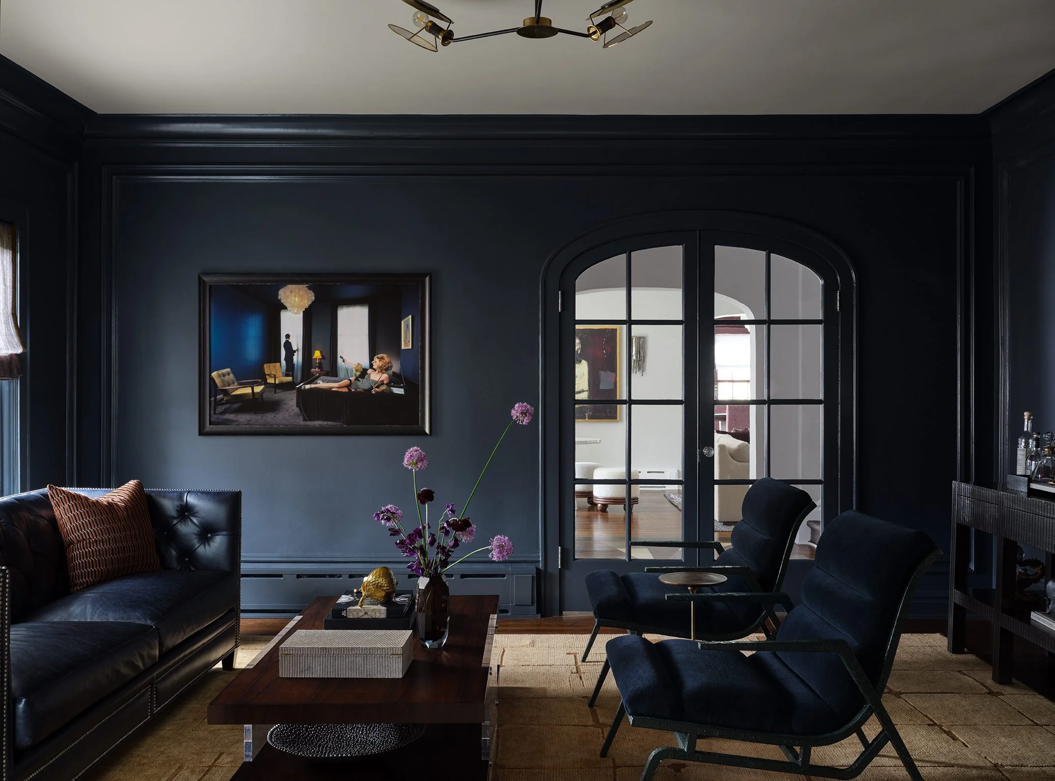 A dark blue living room with black furniture, a wooden coffee table, a mirror, and a vase with pink flowers, with an arched glass door leading to another room with white walls and artwork, and a woman lying on a sofa in the reflection.