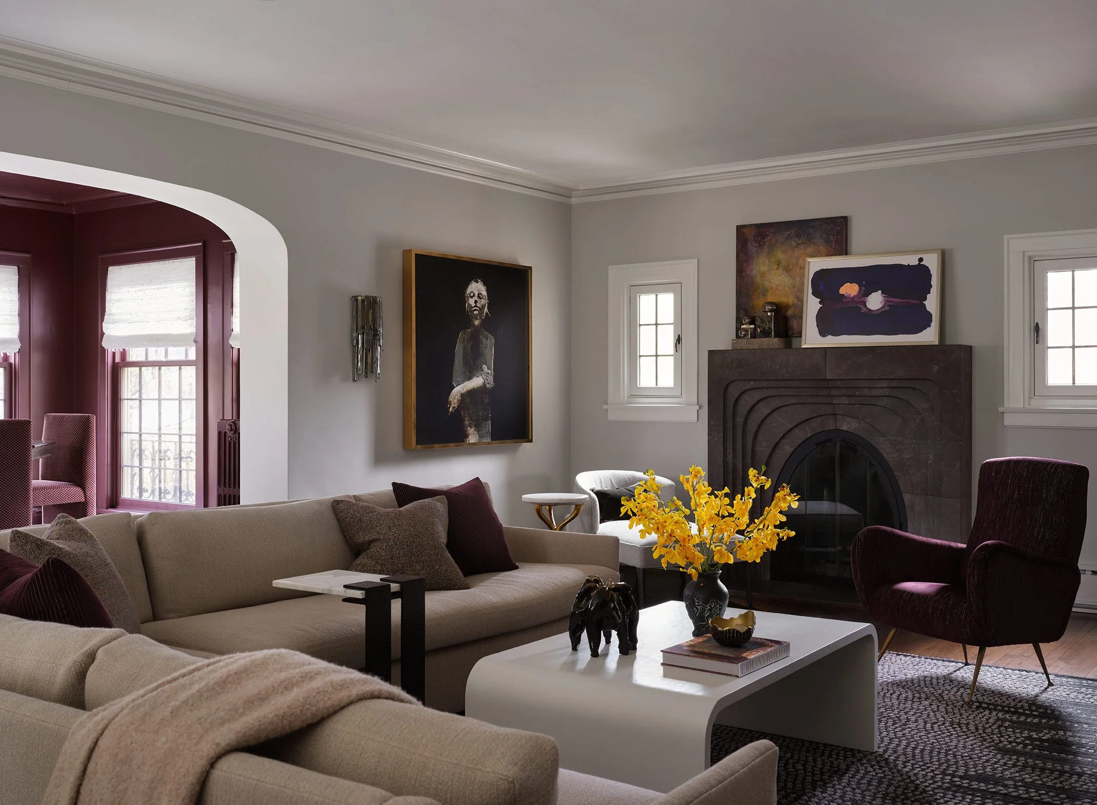 Living room with beige sofa, dark purple armchair, white coffee table with decorative items, yellow flowers in a vase, and wall art including a portrait and abstract paintings. Multiple windows let in natural light.