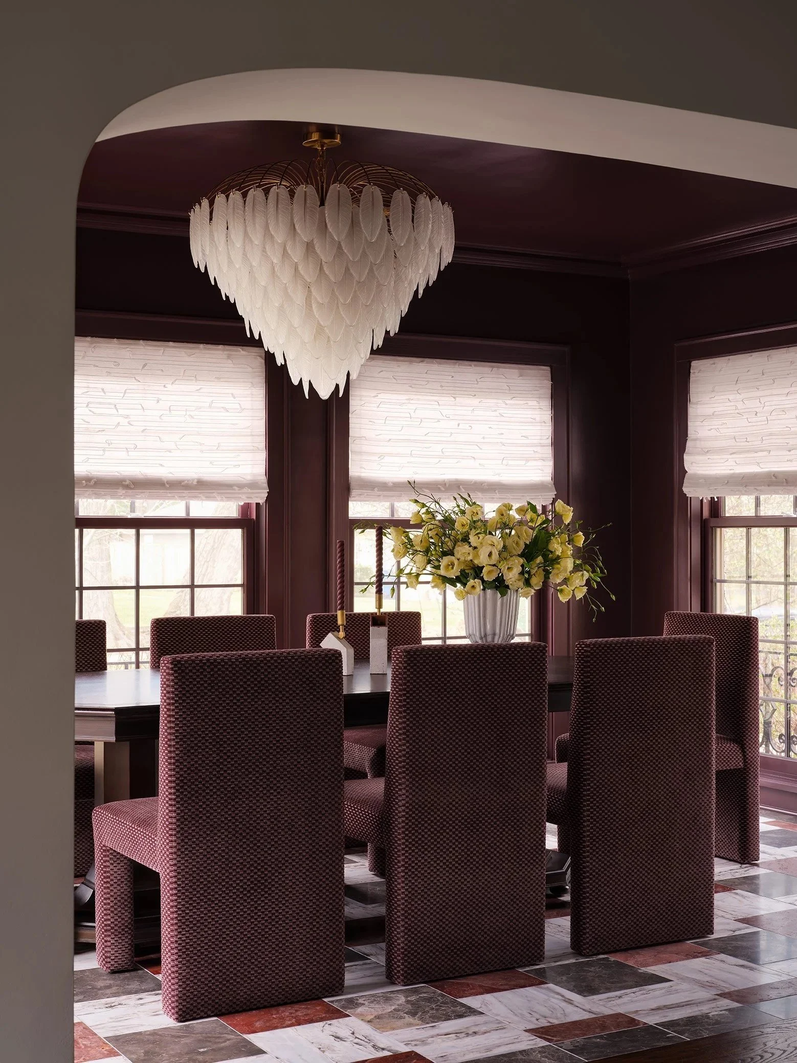 A dining room with a chandelier, a large floral arrangement, and eight upholstered chairs around a table. There are three large windows with white blinds and a dark wood trim.