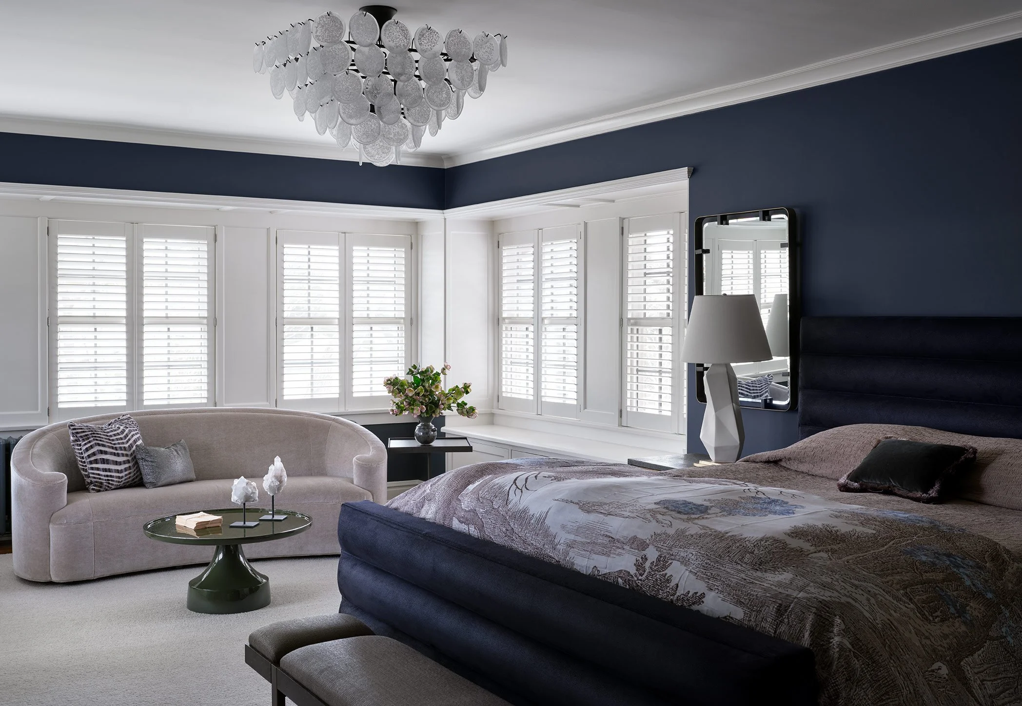 Living room featuring dark blue and white walls, ceiling chandelier, white plantation shutters, beige and dark blue furniture, a mirror, side table with lamp, and decorative flowers.