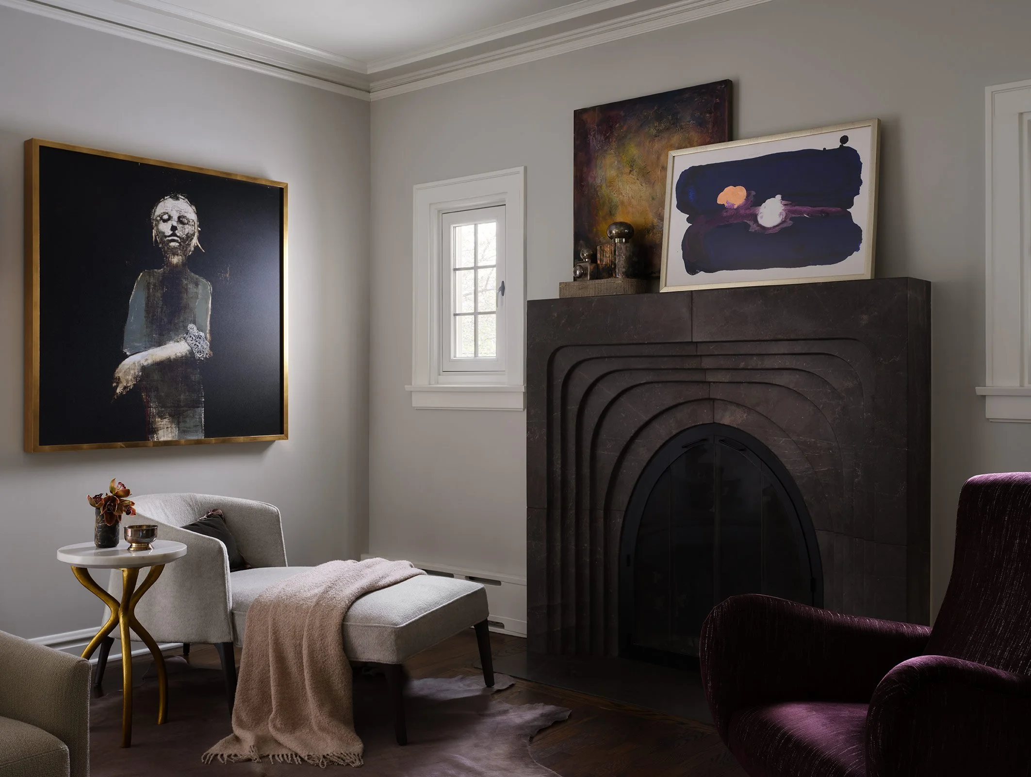 A living room corner with a dark fireplace, abstract paintings on the wall, a gray armchair with a throw blanket, a small round side table with flowers and a candle, and a burgundy armchair.