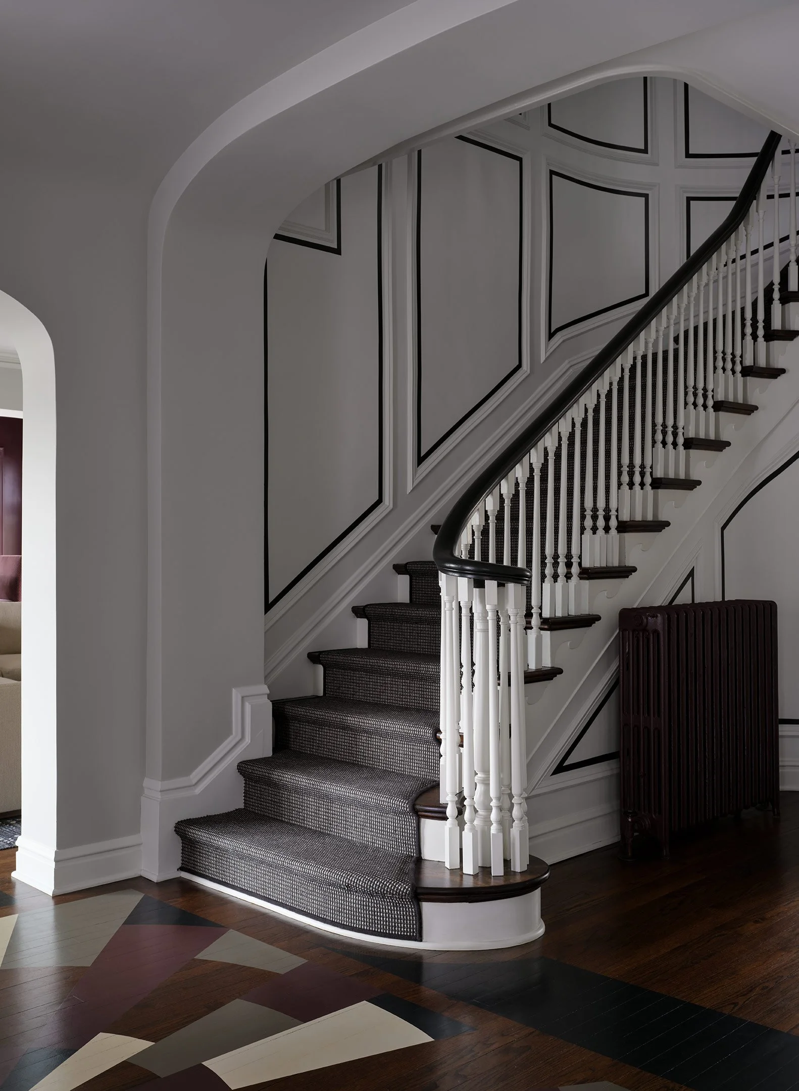 Interior view of a staircase with dark wood handrail and black and white patterned carpeted steps, framed wall paneling, and a geometric-patterned wood floor.