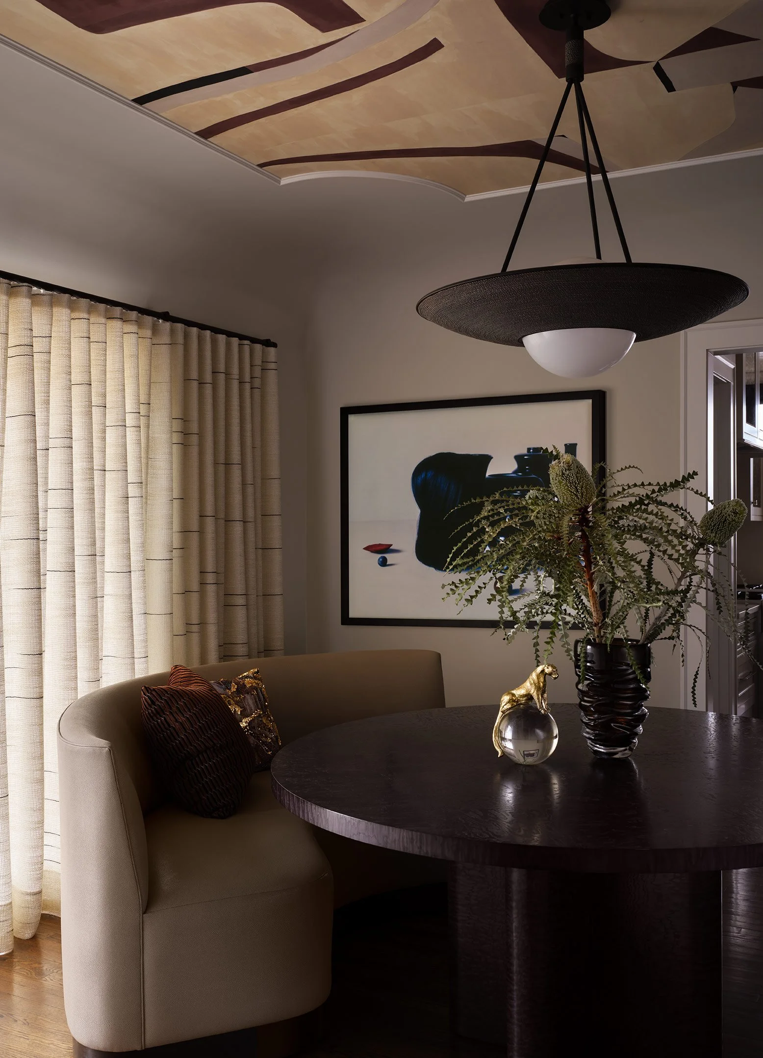 Interior of a living or dining room featuring beige curtains, a framed artwork, a dark round table with decorative items, a beige curved sofa with pillows, and a potted plant. The ceiling has a painted abstract design.