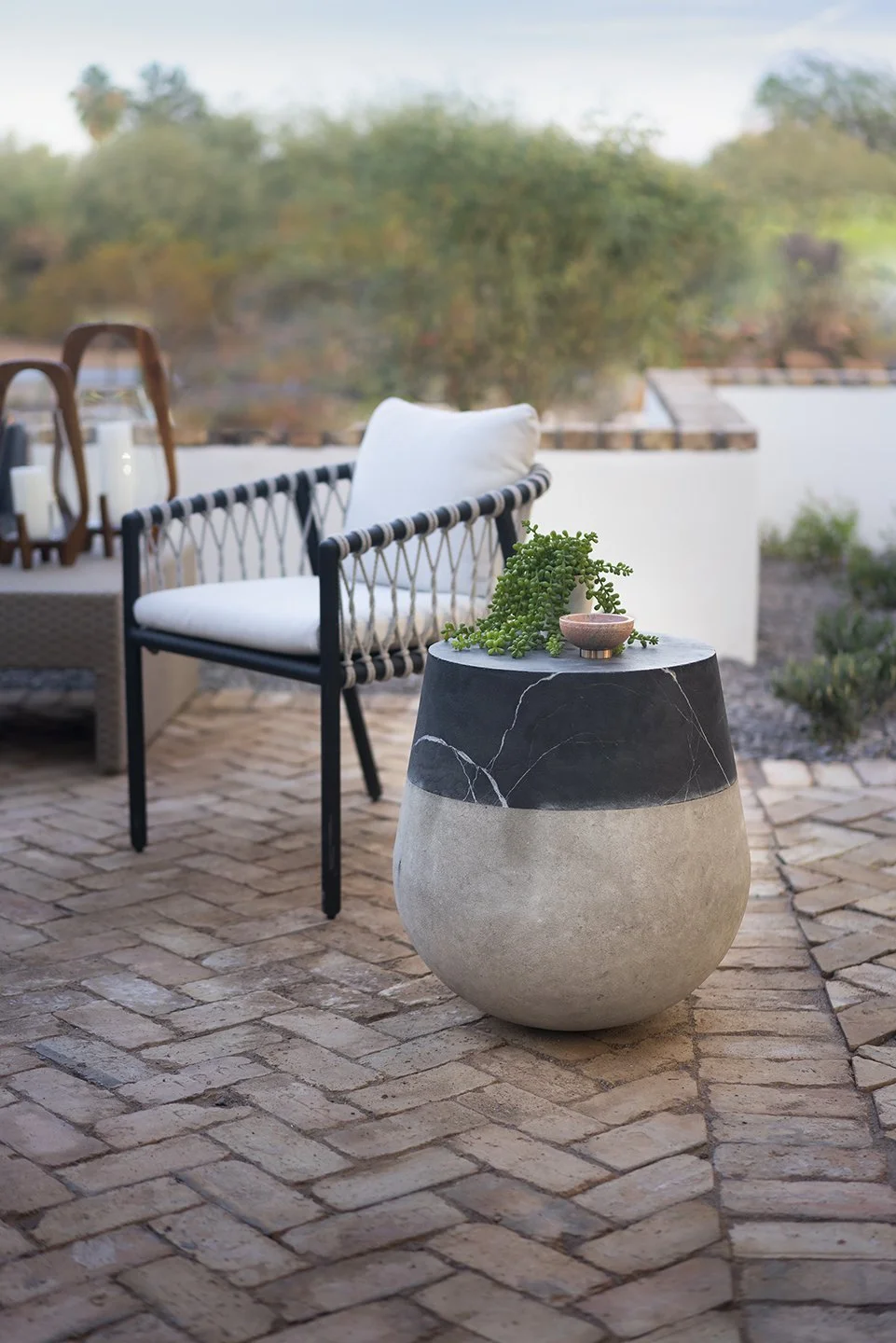 Outdoor patio area with a black and white round stone table, decorated with a green plant and small bowl, and a black framed white cushioned chair on a brick floor, with trees and sky in the background.