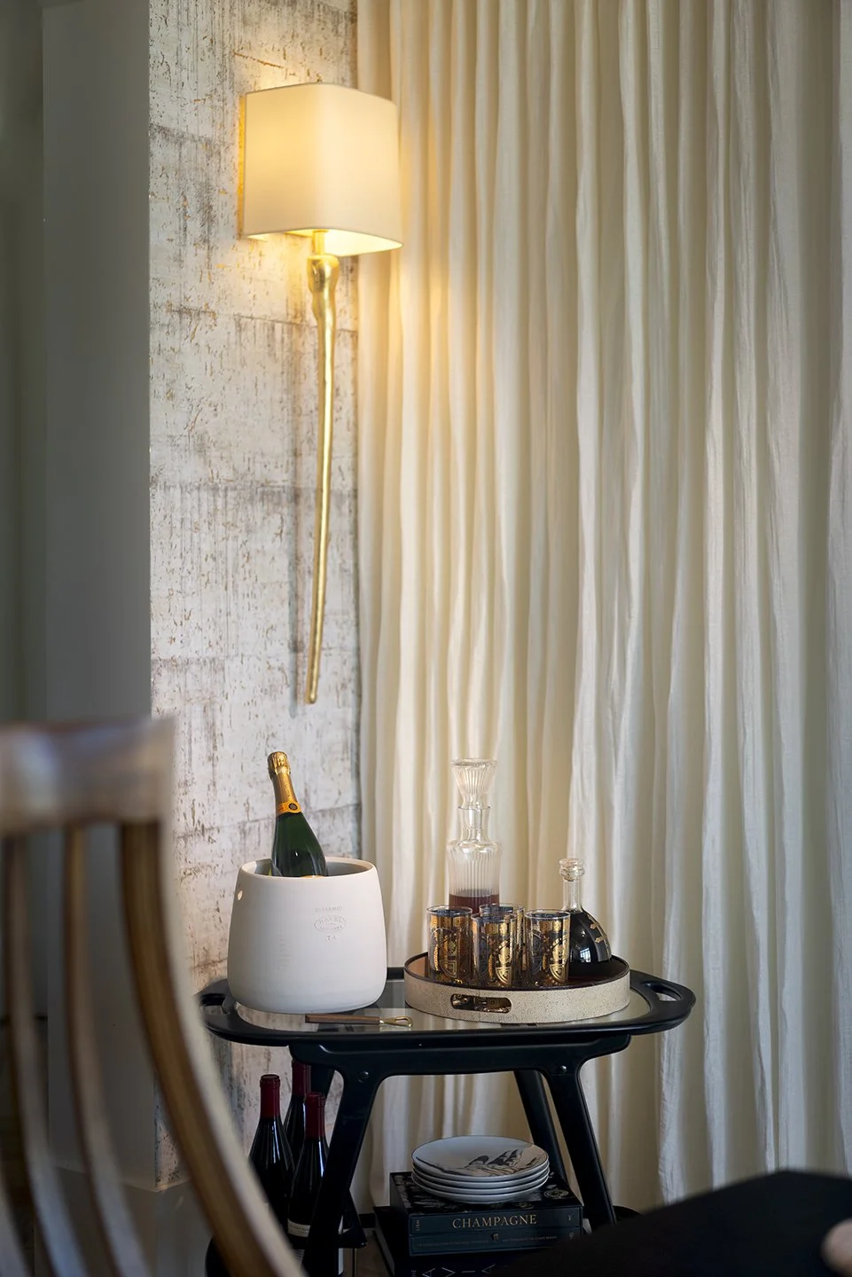 A small round black bar cart with a white ice bucket containing a bottle of champagne, several glasses, a decanter, and a black vintage-style telephone. Behind the cart, there are cream curtains and a textured wall with a wall-mounted lamp emitting w