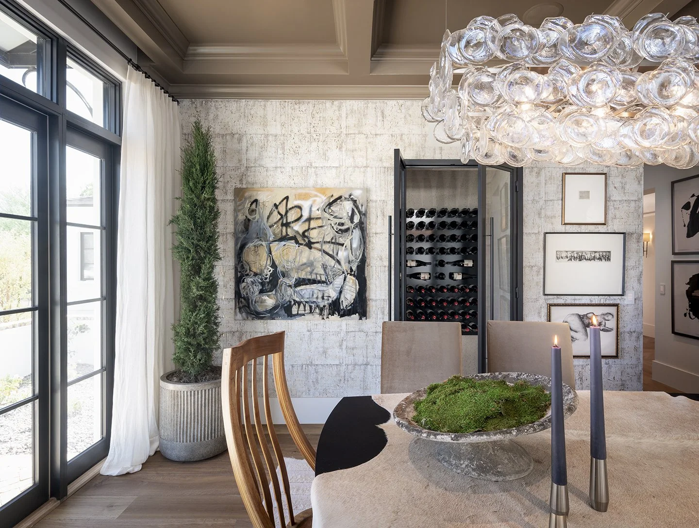 Modern dining room with glass chandelier, abstract art on white brick wall, potted evergreen plant, and wine cooler with bottles, along with framed artwork on the wall near open doorway.