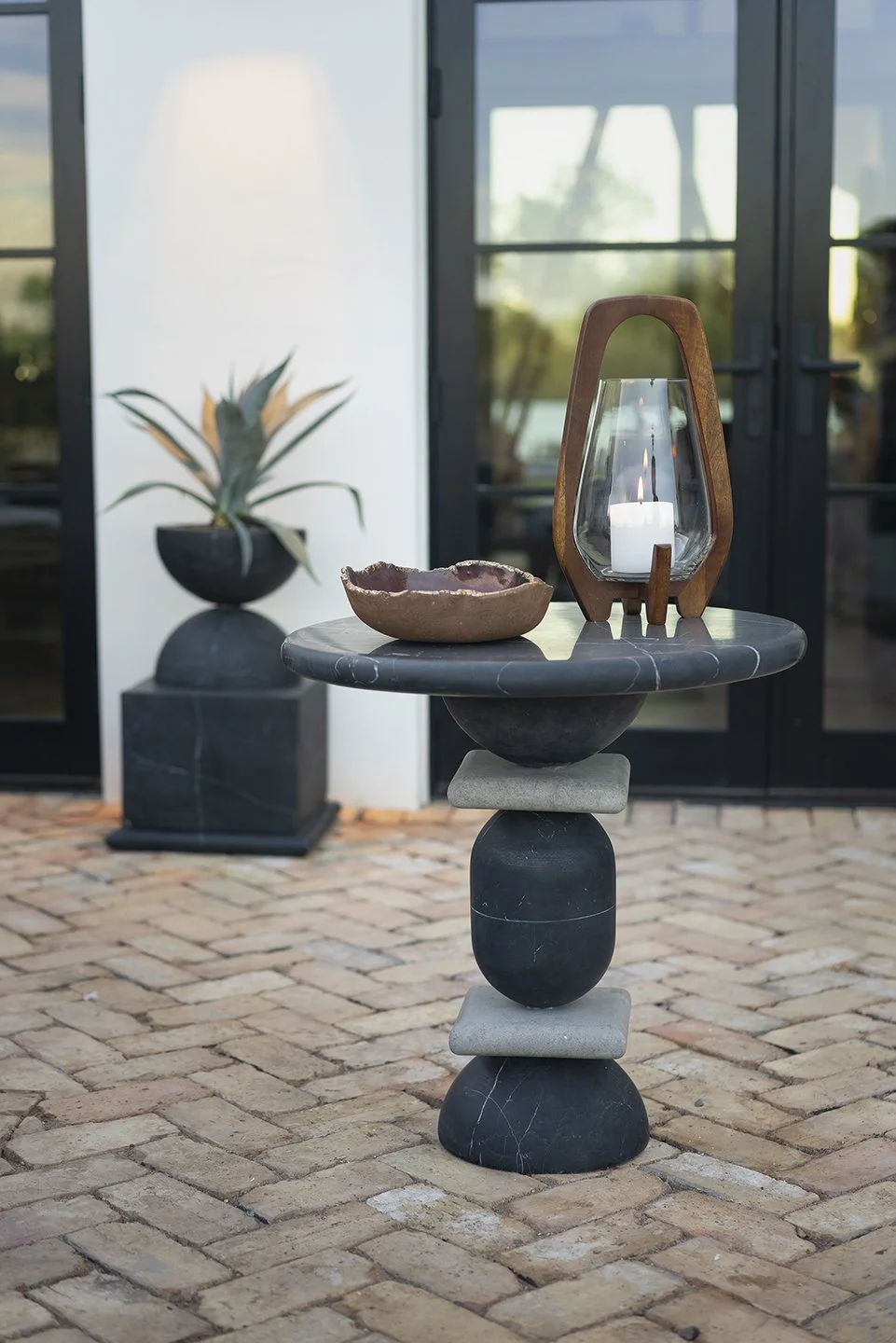 Decorative table with a candle inside a glass lantern, a ceramic bowl, and a stone on the bottom, outside on a brick patio with a potted plant in the background.
