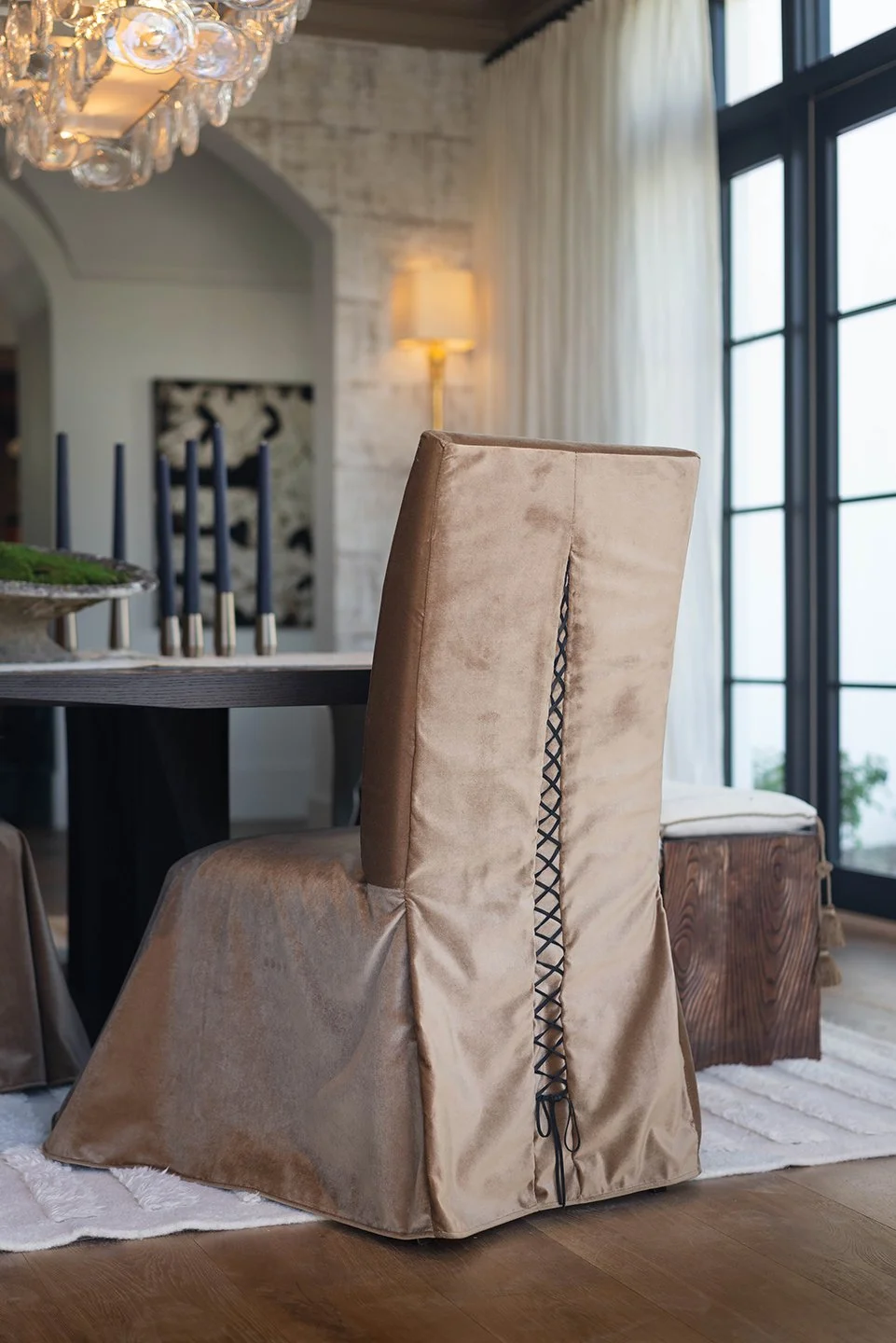A beige upholstered dining chair with a back lace-up detail sitting on a white shag rug in a modern dining room with a glass door, curtains, and a large chandelier.