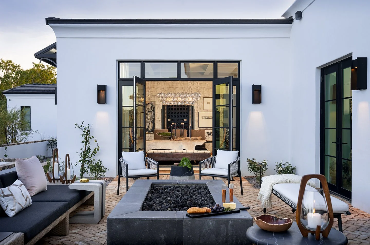 Outdoor patio with black and white seating, a fire pit, candles, and decorative items, overlooking a modern house with large black-framed windows and glass doors.