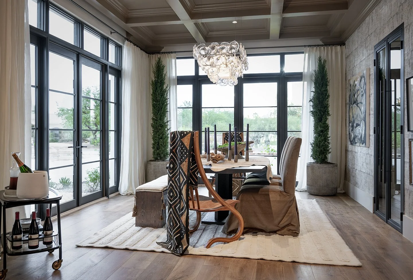 A well-lit dining room with large windows and glass doors showing an outdoor view. The room has a dining table with chairs, a chandelier, potted plants, and decorative artwork on the wall.