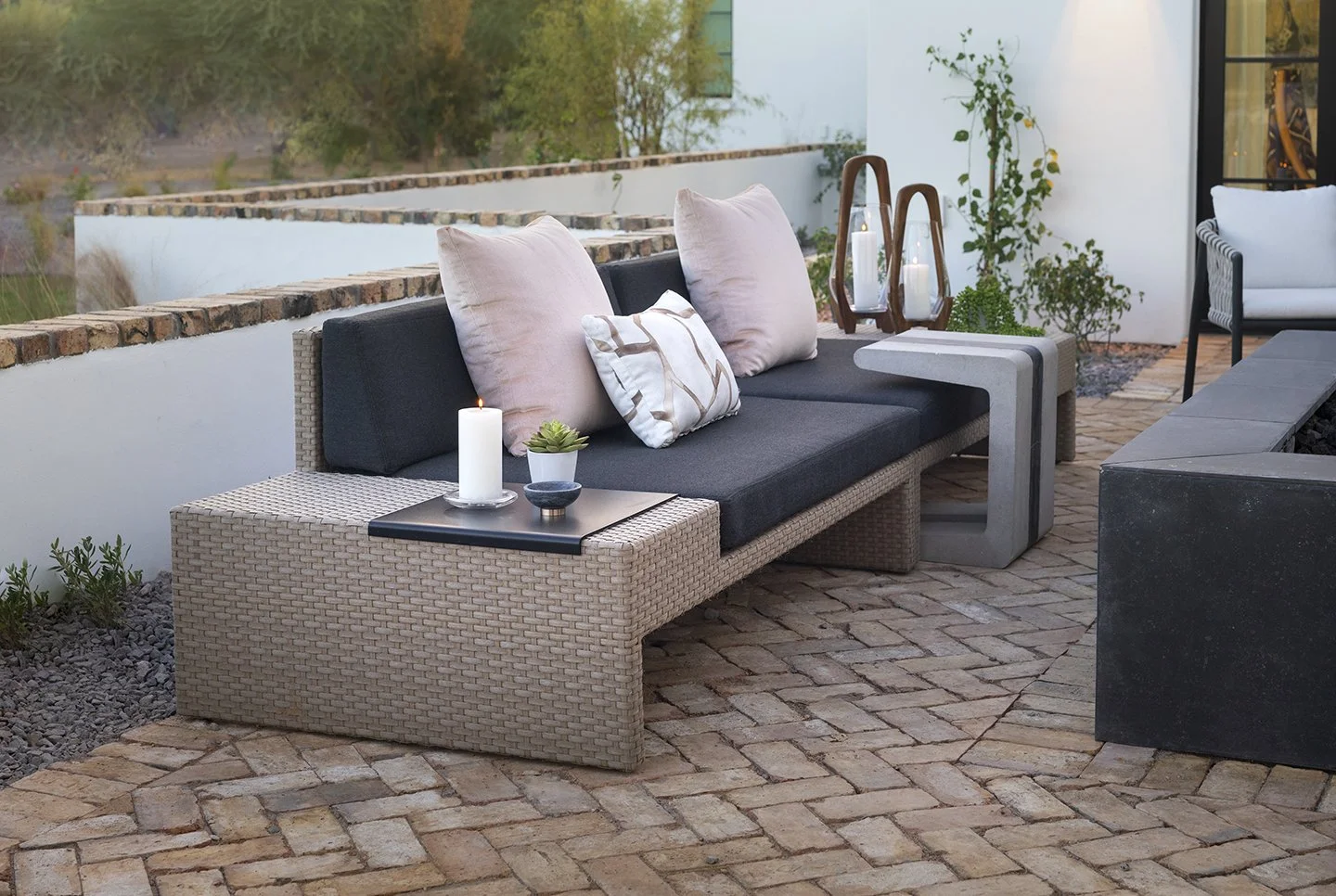 Outdoor patio with a woven wicker sofa with black cushions and pink pillows, a small black table with candles and plants, concrete side tables, potted plants, and a brick patio with a white wall and view of trees.