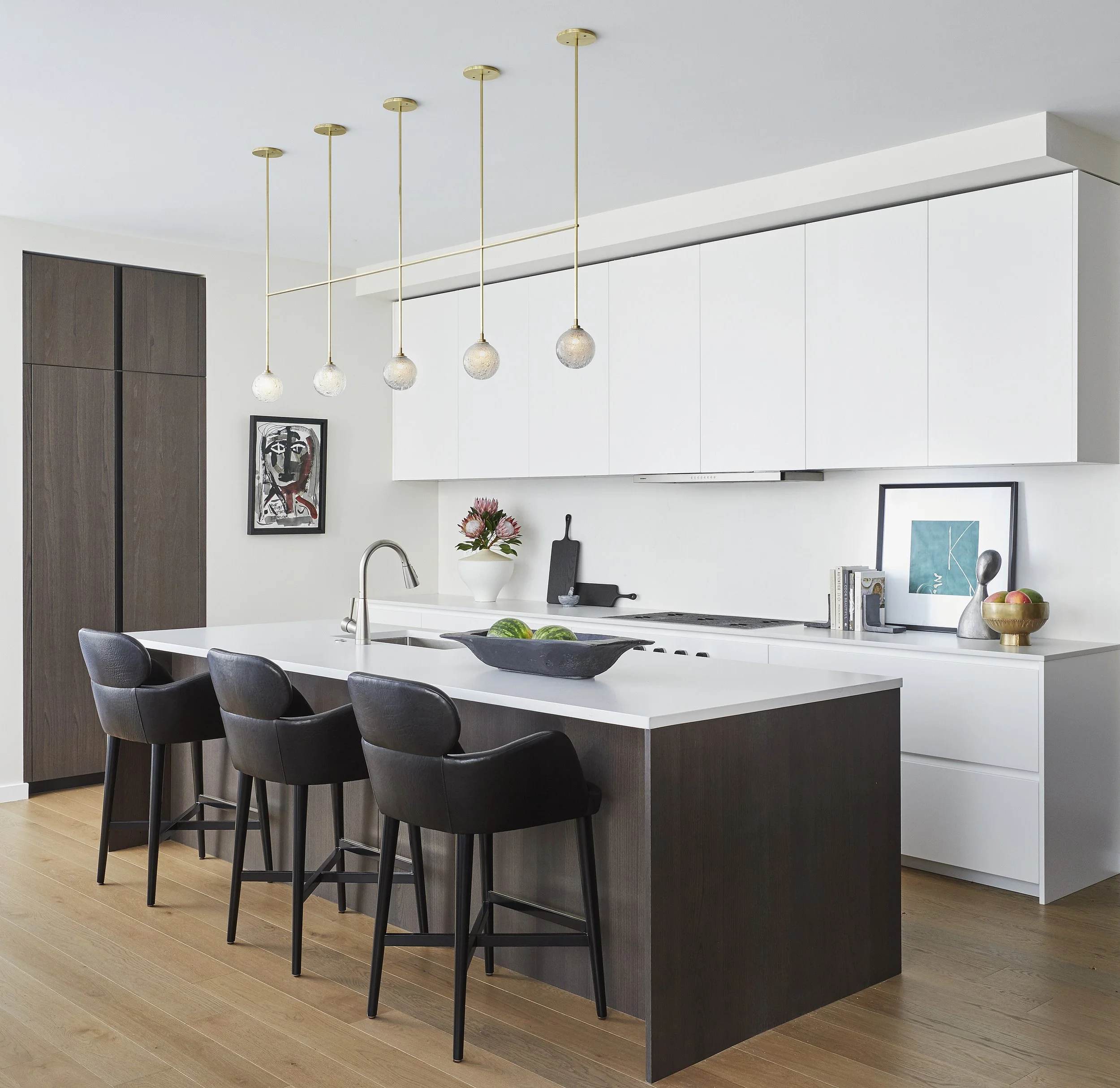 Modern kitchen island with dark wood base, white countertop, black chairs around it, and pendant lights hanging above. Kitchen decor includes a vase of pink flowers, framed artwork, books, and a bowl of apples. The background features a built-in cabi