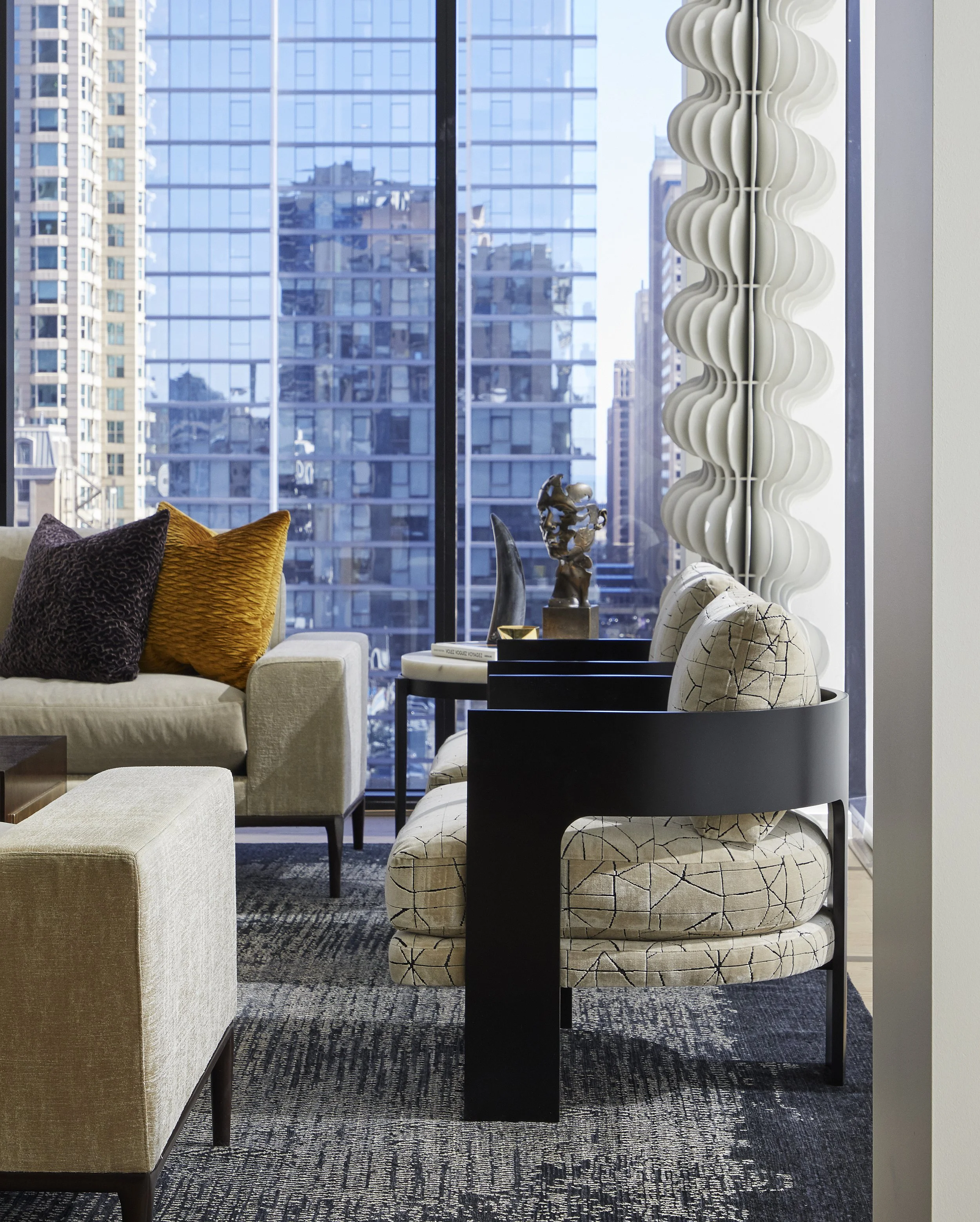 Modern living room with a beige sofa, decorative cushions, and a black coffee table. Tall sculptural white floor lamp and cityscape view through large glass windows.