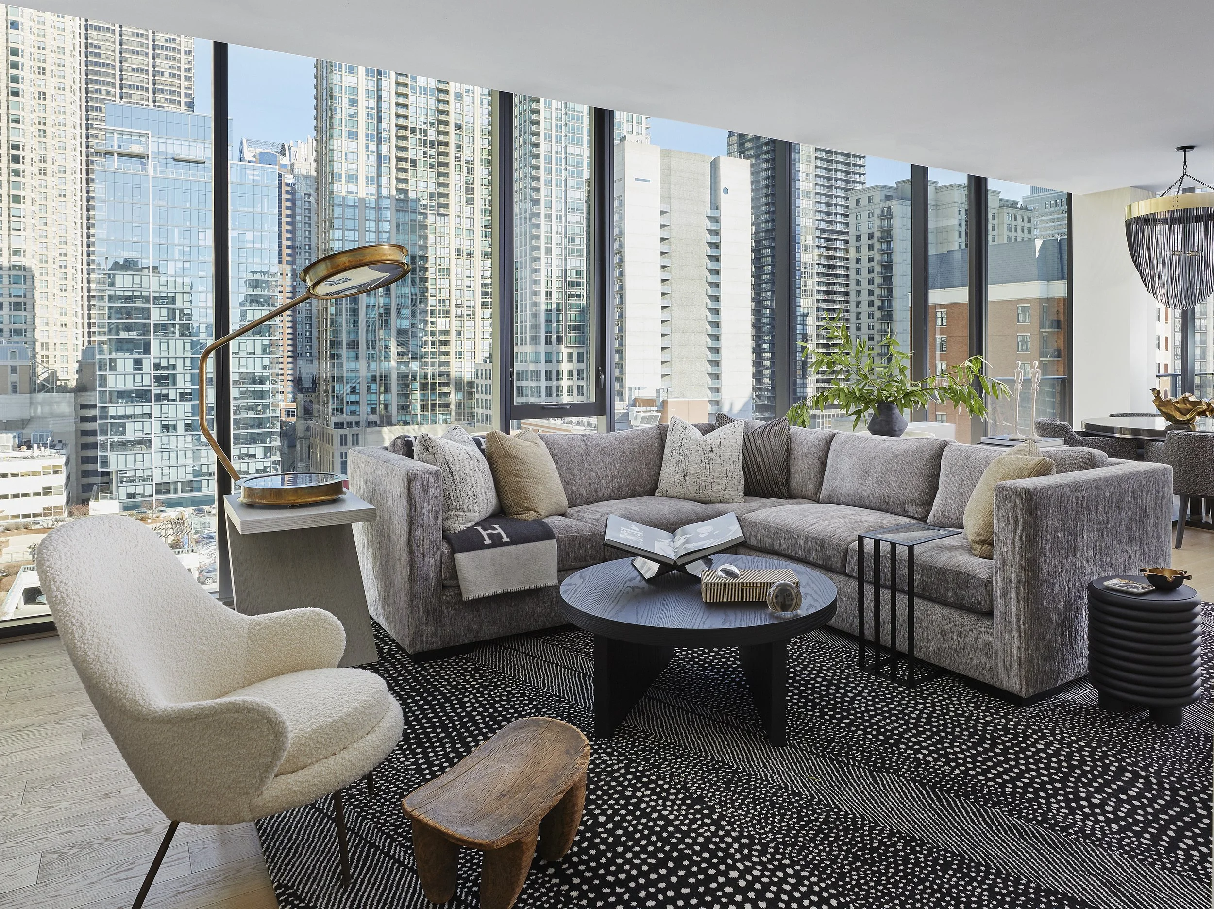 Modern living room with large windows showing a city skyline, beige sectional sofa, white armchair, round black coffee table, black and white patterned rug, and decorative plants and sculptures.