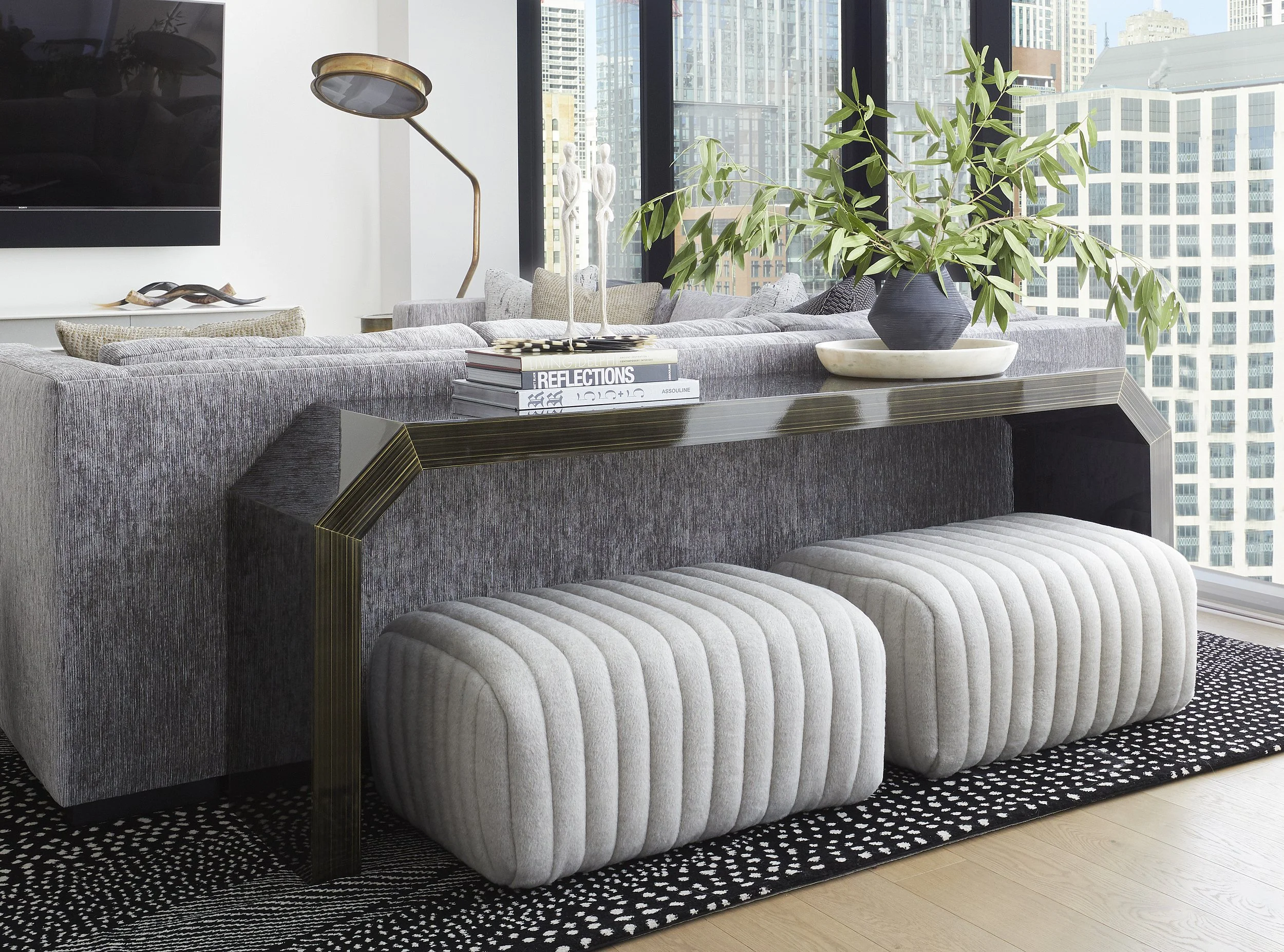 Modern living room with gray textured sofa, white quilted ottoman, black and white patterned rug, large black vase with green leafy plant, coffee table with books, and city skyline visible through large floor-to-ceiling windows.