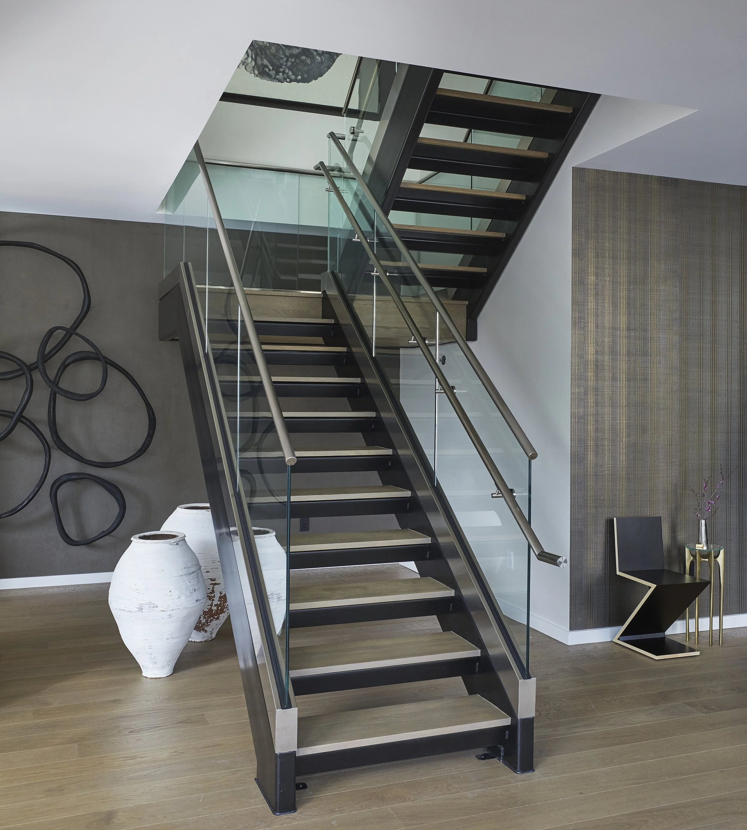 Modern staircase with glass sides and metal railings, flanked by decorative white vases and black art pieces, in a contemporary interior.