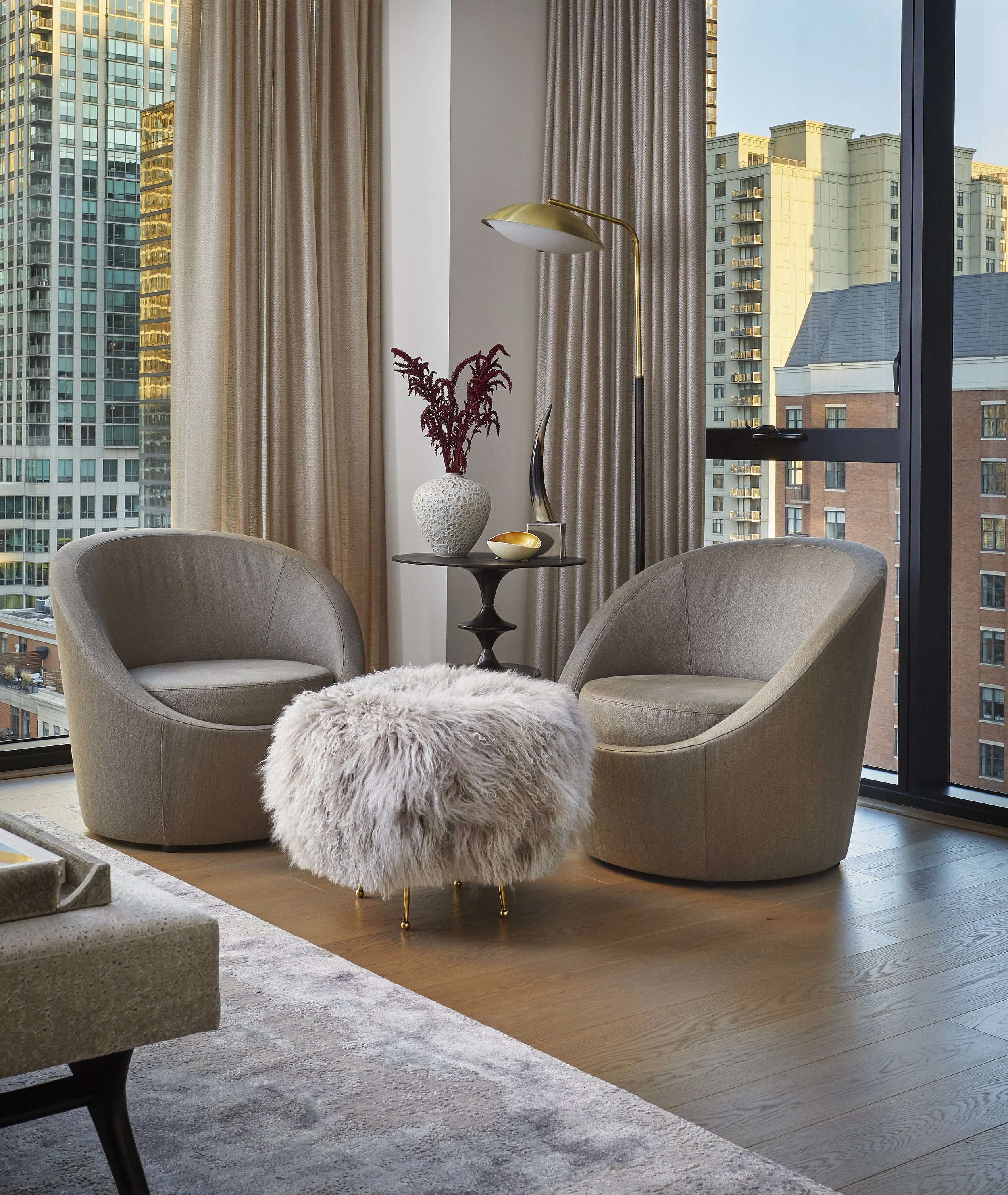 Living room with large windows showing city buildings, beige curtains, a round side table with a decorative vase and a curved statue, two beige armchairs, a fluffy white stool, a gold and white table lamp, and a mix of hardwood and carpeted flooring.