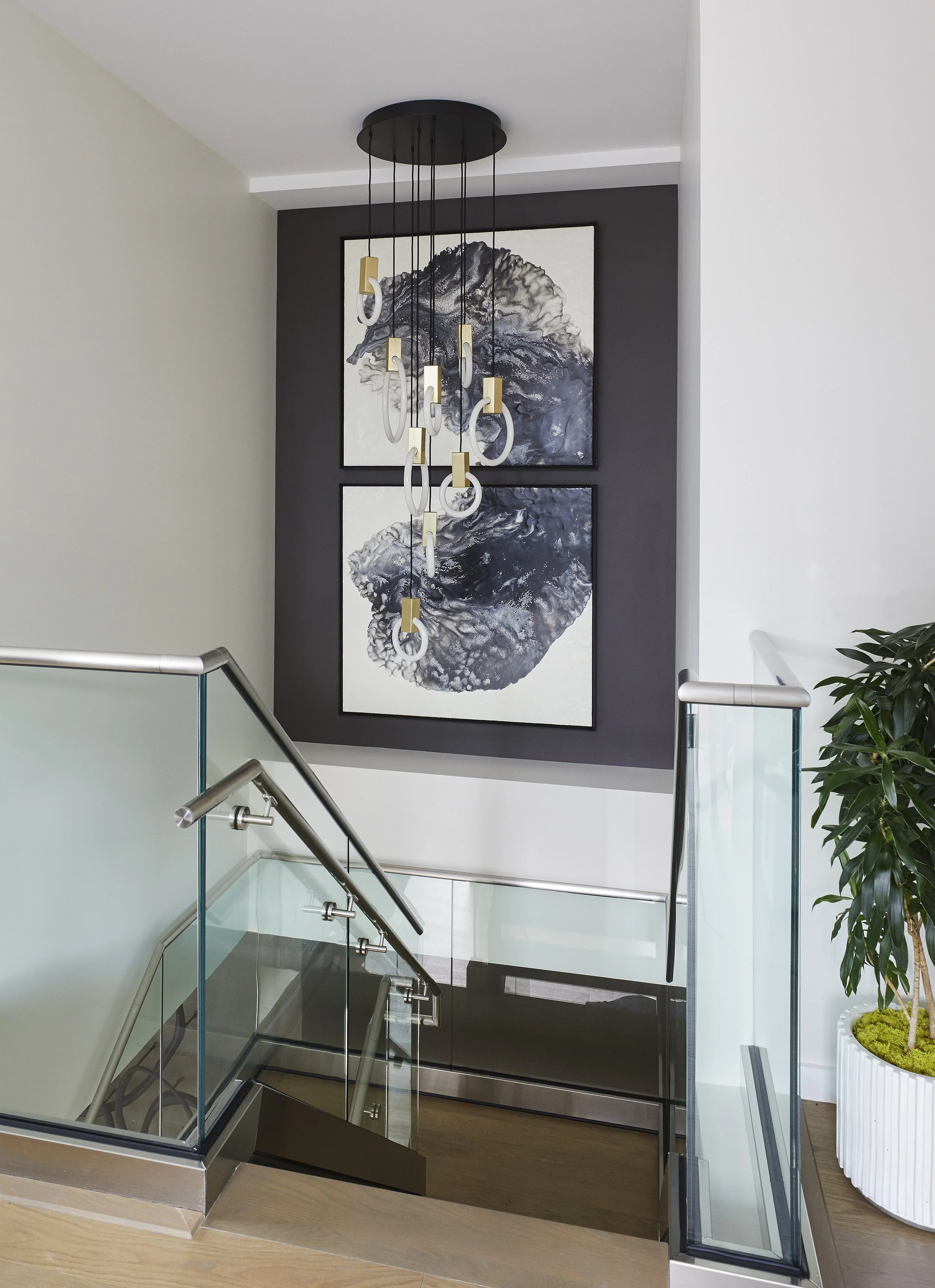 Modern stairway with glass railing, black stairs, white and gray walls, two large abstract black-and-white paintings, hanging light fixture with white and gold details, potted green plant on white stand.