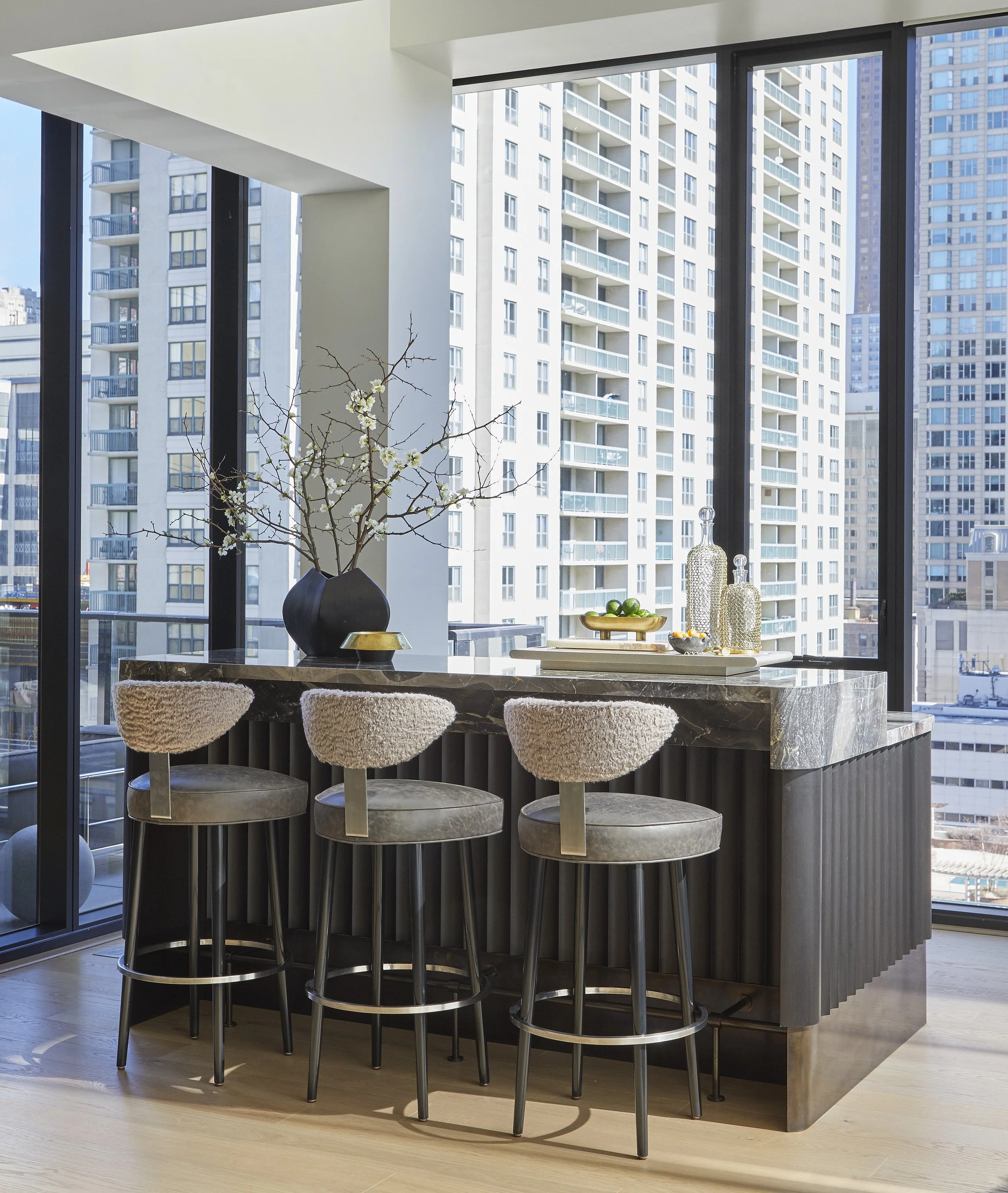 Modern kitchen island with a marble surface and four barstools with plush, light-colored upholstery, in front of large floor-to-ceiling windows showing tall city buildings.